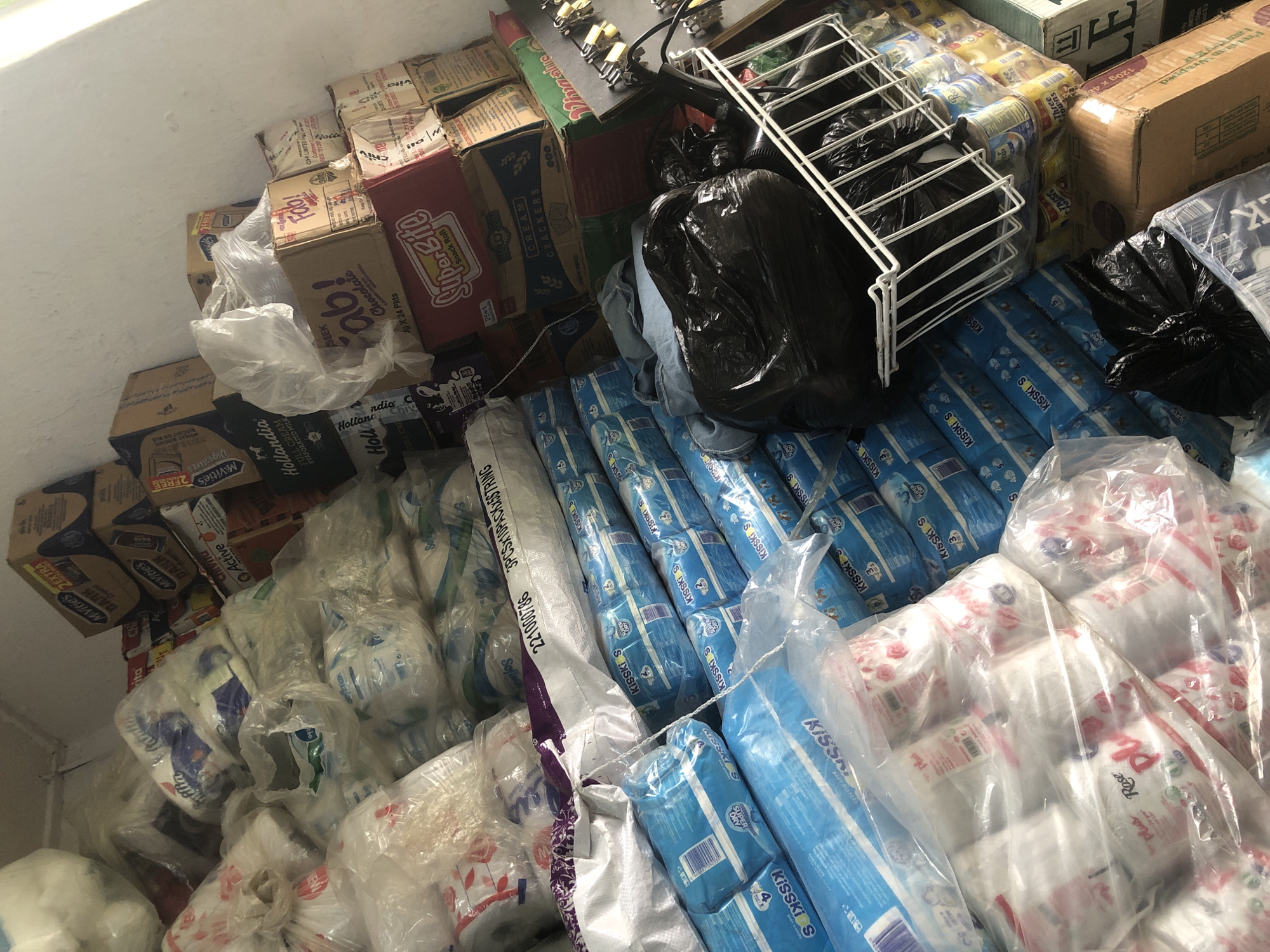 This is a high-angle, close-up shot of a cluttered storage space in Ado, Nigeria, filled with various goods. The items are stacked haphazardly, creating a chaotic yet organized appearance. Dominating the scene are rows of blue-packaged baby diapers, labeled "KISSKis S." Next to these are packages of toilet paper with floral patterns.
Further back, several cardboard boxes are visible, some with brand names like "McVities," "Hollandia," and "Fab! Chocolate." One box is labeled "SuperBite Snack Roll." There are also cans of what appear to be evaporated milk.
In the foreground, a white wire basket holds a black garbage bag filled with what seems to be plastic sheeting or fabric. Another black garbage bag lies on the floor to the left of the basket. The environment appears to be an indoor storage area, possibly a storeroom or a shop's back room, with plain, light-colored walls. The overall impression is of a busy, possibly commercial, environment where inventory is being stored.