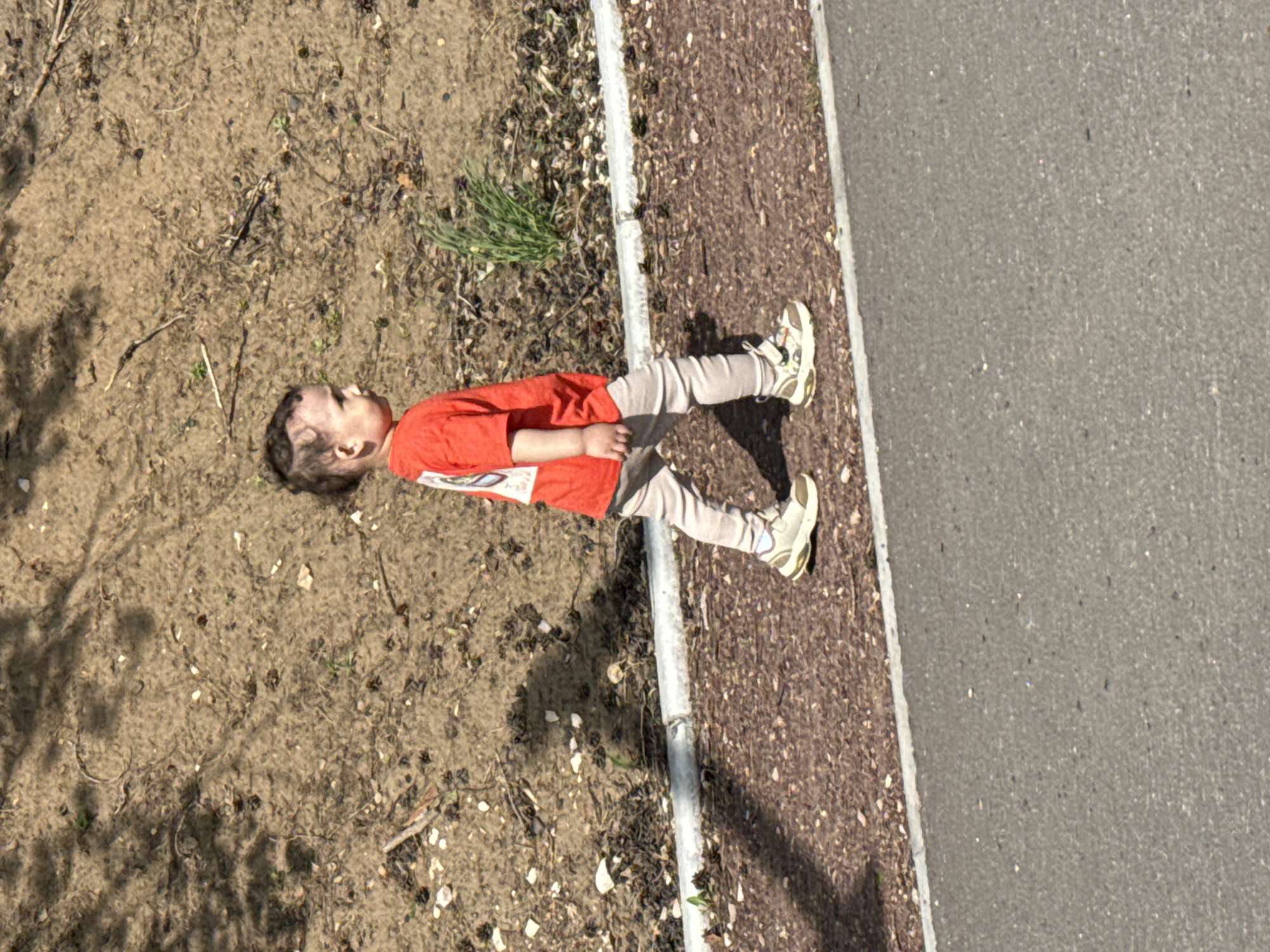 A child lies on their back near a sidewalk bordering a dirt area in what seems to be an urban setting. The child is wearing a red shirt, light grey leggings, and sneakers. Part of the sidewalk is paved in brown and part is grey asphalt. The dirt area has sparse vegetation. The location is likely Chaikovsky, Russia.