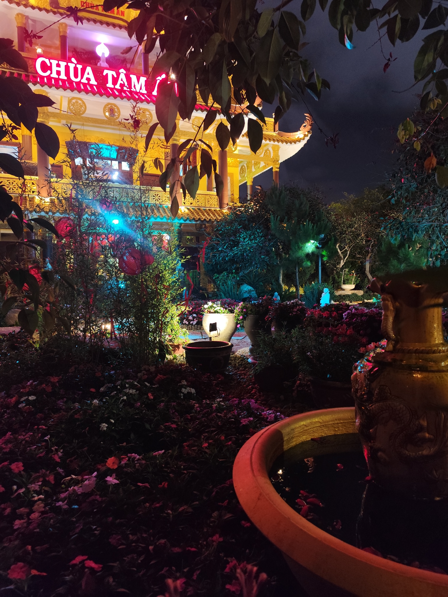 The image captures a night scene in Da Lat, Vietnam, showcasing the Chua Tam Lam temple illuminated with neon lights. The temple, a multi-tiered structure, dominates the background, adorned with red neon signs reading "Chua Tam Lam." In the foreground, a garden features potted plants, flowers, and a decorative water fountain with a yellow hue. The lighting is a mix of artificial colors, creating a vibrant yet slightly dark atmosphere. No people are visible in the shot. The setting suggests a blend of cultural architecture and cultivated nature, indicative of the temple's grounds at night.