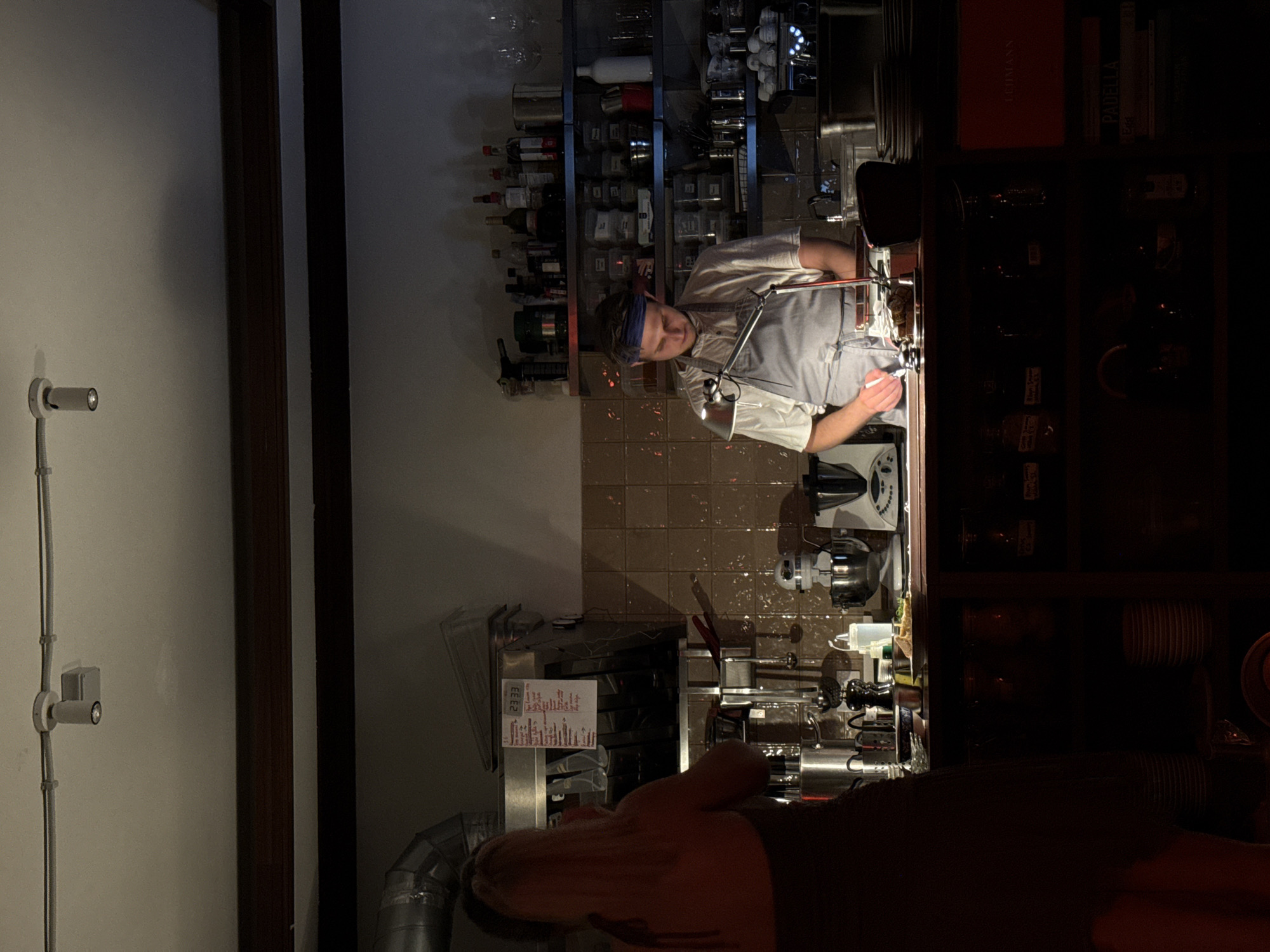 This indoor image, captured in Belgrade, Serbia, shows a man working behind a counter in what appears to be a bar, restaurant, or high-end kitchen, set during the evening or night as indicated by the dim ambient lighting and prominent use of task lights.
The main subject is a man wearing a light-colored shirt or chef's jacket, a dark apron or bib, and a dark headband. He is positioned at a counter, intently focused on an object under the bright illumination of a metallic task lamp. Various kitchen appliances and tools, including a blender or mixer and other stainless steel equipment, are arranged on the counter around him.
Behind the man, shelves are stocked with numerous bottles, likely spirits or wine, alongside an assortment of jars and containers. The wall behind these shelves is covered with brown, square tiles. To the man's right, a dark wooden shelving unit holds additional bottles and several books. Visible on the book spines are the titles "LEHMANN" (on a red book) and "PADELLA."
In the foreground, the arm and shoulder of another person are partially visible, suggesting a customer or colleague is present, possibly observing the activity. To the far left, a white wall features a simple spotlight fixture and exposed electrical conduit. On a metal surface near the man, a white paper with the numbers "23 33" and some illegible text is attached. A large metallic ventilation pipe is also visible in the lower-left portion of the frame. The scene conveys a focused, professional atmosphere.
