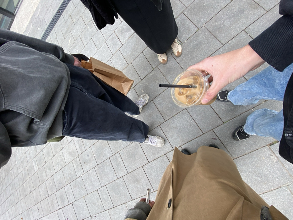 An overhead-angle shot captures four individuals standing on a light grey tiled pavement in an urban area of Tel-Aviv, Israel.
In the immediate foreground, a person's left hand holds a clear plastic cup filled with iced coffee, containing a black straw and ice cubes. This person wears light blue wide-leg jeans and black sneakers. To their left, another individual wears a long, tan-colored trench coat and black shoes; their left hand is visible holding a silver-colored smartphone with a dark screen. Directly opposite the foreground person, at the top center of the frame, stands an individual wearing a dark grey, faded hooded sweatshirt over a green garment, dark grey wide-leg jeans, and white athletic shoes. This person's right hand holds a brown paper shopping bag. To the right, partially visible, a fourth individual wears black wide-leg trousers or a skirt and light brown and white athletic shoes.
The ground consists of square light grey paving stones arranged in a grid pattern. A narrow metal drainage grate is visible along the upper right edge of the paved surface, bordering a dark, undefined structure. All individuals appear to be standing still, facing generally towards the camera's point of view.