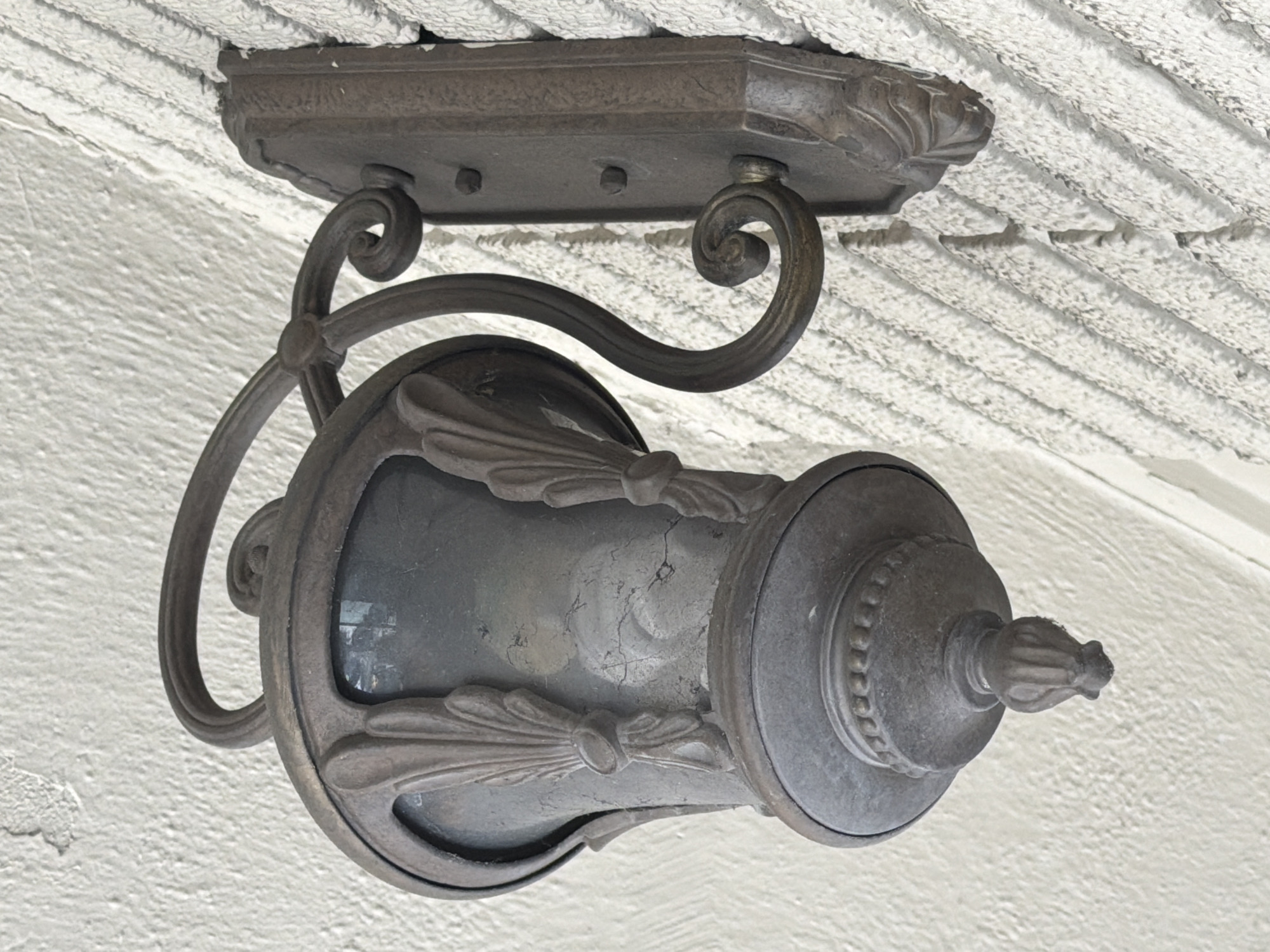 An outdoor wall-mounted lantern with a dark metal finish is affixed to a textured white wall. The lantern has a decorative, bell-shaped body with glass panels and ornate metalwork that resembles leaves. The base of the lantern tapers to a decorative finial. The mounting bracket is also dark metal and features scrolling details. The wall has a rough, stucco-like texture with prominent horizontal ridges. The scene appears to be well-lit, suggesting daylight.