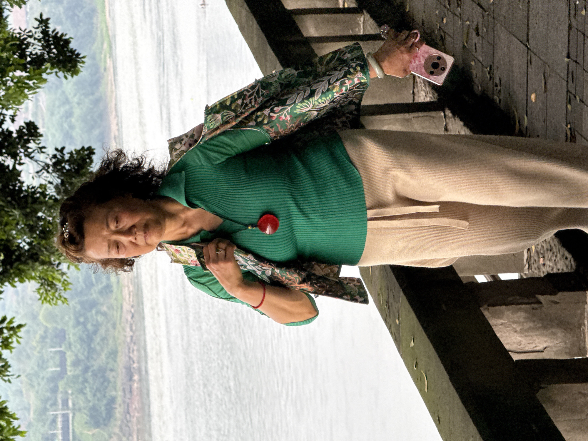 An older Asian woman stands outdoors, holding a pink smartphone in her right hand, while facing a broad expanse of water, likely a river. She wears a green, collared, ribbed shirt with a large red pendant, beige pants, and a floral-patterned jacket. She stands on a wooden structure that seems to be a bridge or elevated walkway. The river is the backdrop, stretching out behind her, with trees lining the far shore, partially obscured by mist or haze. The photograph appears to have been taken near the river in Jiangjin, China.