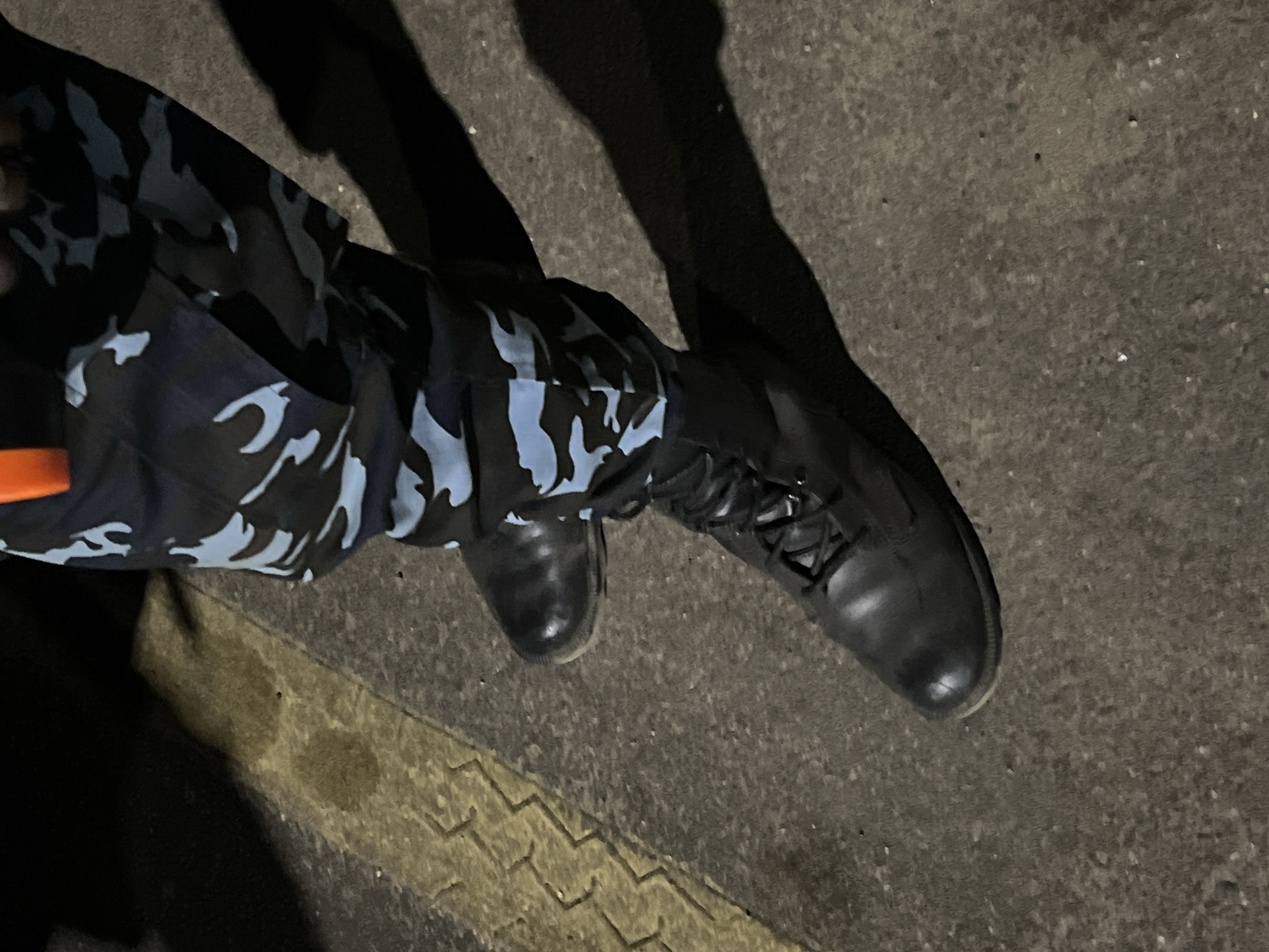 The image shows a person's legs and feet on a concrete surface. The person is wearing black combat boots and camouflage pants with a black and light blue flame pattern. The background is a concrete surface with a yellow textured line. The shadows suggest the scene is at night with artificial lighting. It is not possible to determine the exact location or any specific actions taking place from the image alone, and Nigeria/Dam Camp cannot be verified.