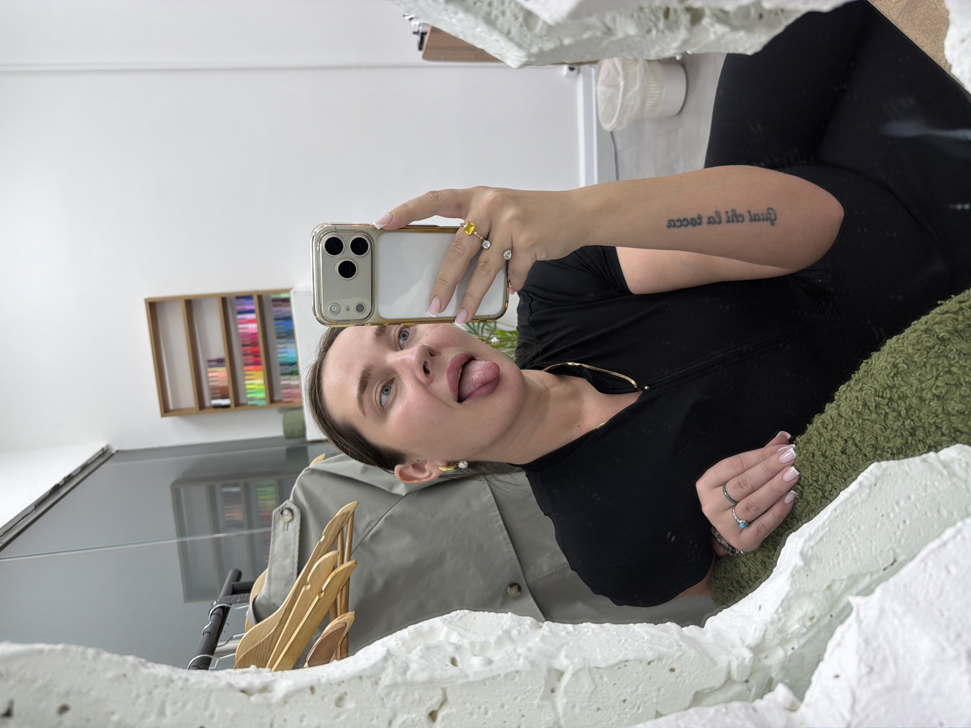 The photograph captures a woman taking a selfie in front of a mirror. She is holding an iPhone with a distinctive triple-lens camera system, encased in a gold-toned case. The woman is wearing black clothing and jewelry, including rings and earrings. She has a tattoo on her upper arm. The reflection in the mirror shows elements of the environment, including a shelf with what appears to be a collection of colored pencils or art supplies, a gray coat, and some wooden items. The setting is an interior space with white walls. There is no clear information to indicate the location of this picture in Moscow, Russia.