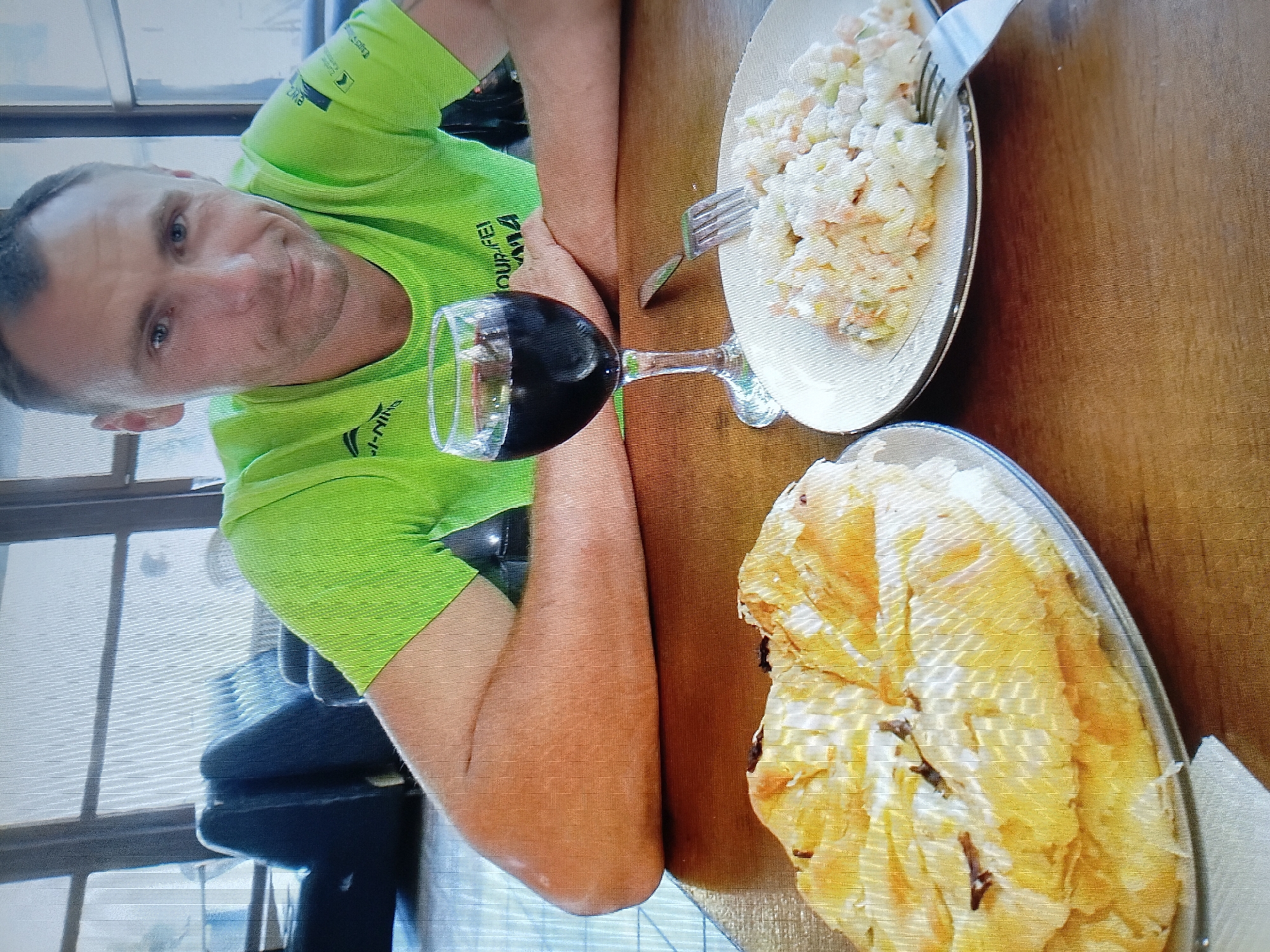 A man with short brown hair and blue eyes is seated at a wooden table, wearing a neon green t-shirt with logos. In front of him, a glass of red wine and two plates of food are visible: one with a large, golden-brown pastry and another with a salad. The background shows a seating area with dark furniture and large windows. The scene appears to be indoors, possibly a restaurant or cafe. It is not possible to determine the location within Tiraspol, Moldova from the visual information provided.
