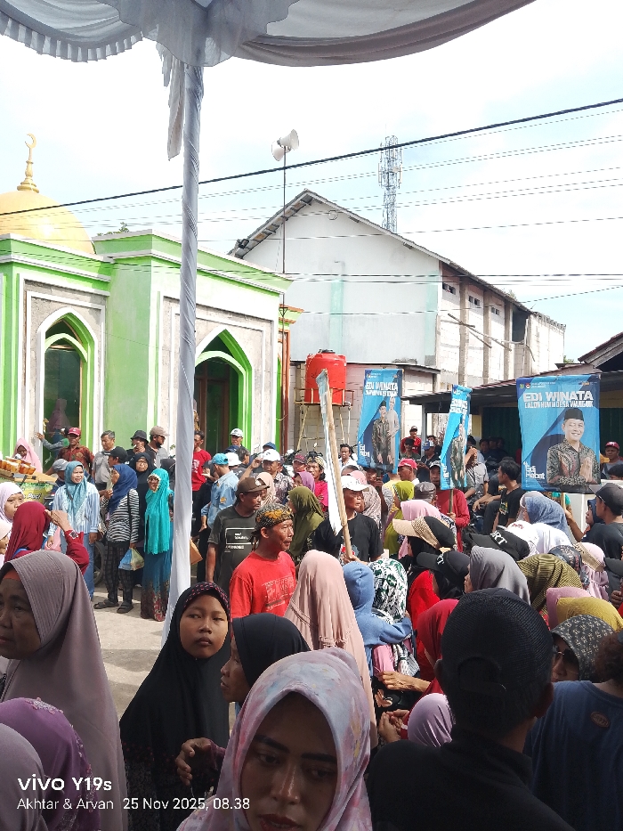 A vibrant gathering of people is assembled outdoors, with a mosque featuring a green and white facade and a golden dome in the background. The crowd is diverse, with many women wearing hijabs. Banners display the image of "EDI WINATA," likely a candidate, with accompanying text. A water tower and utility pole stand behind the crowd. The photo's timestamp indicates "25 Nov 2025, 08.38," and a "vivo Y19s" watermark is visible.