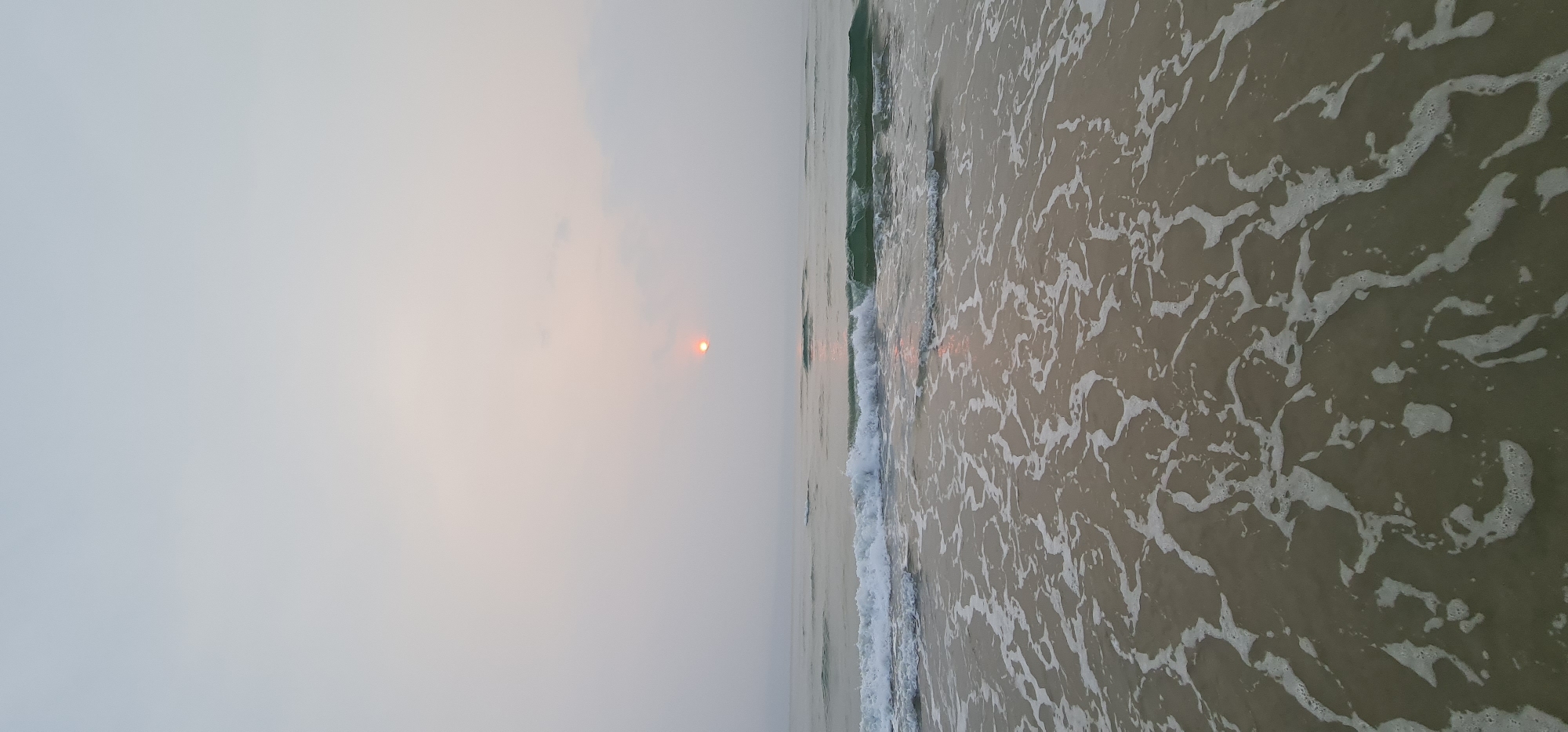 The image shows a coastal scene in Đà Nẵng, Vietnam, at sunrise or sunset. The sun is visible through a hazy sky. Gentle waves are breaking on the sandy shore. The water is a light tan color, with white foam patterns created by the wave action. There are no people visible in the frame.