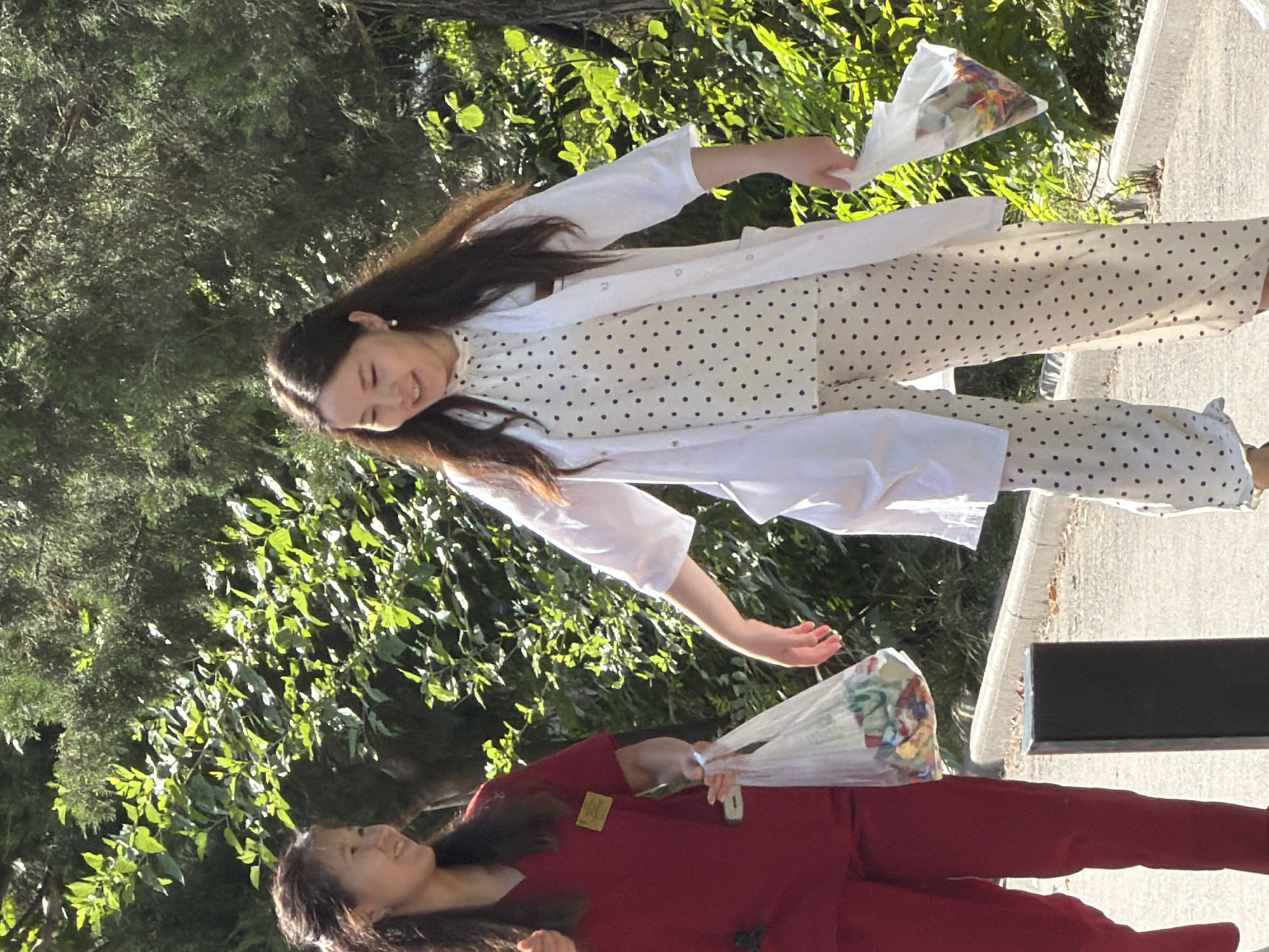 Two Asian women are walking outdoors in a park-like setting. The woman on the left wears a red scrub top and red pants, and carries a clear plastic bag filled with colorful items. She has dark hair pulled back and is smiling. The woman on the right wears a white polka-dot top and matching pants, with a white lab coat over her. She also carries a clear plastic bag filled with colorful items and is smiling and looking down. They are walking on a paved path next to a curb, with lush green foliage in the background. A dark rectangular object is visible in the foreground.