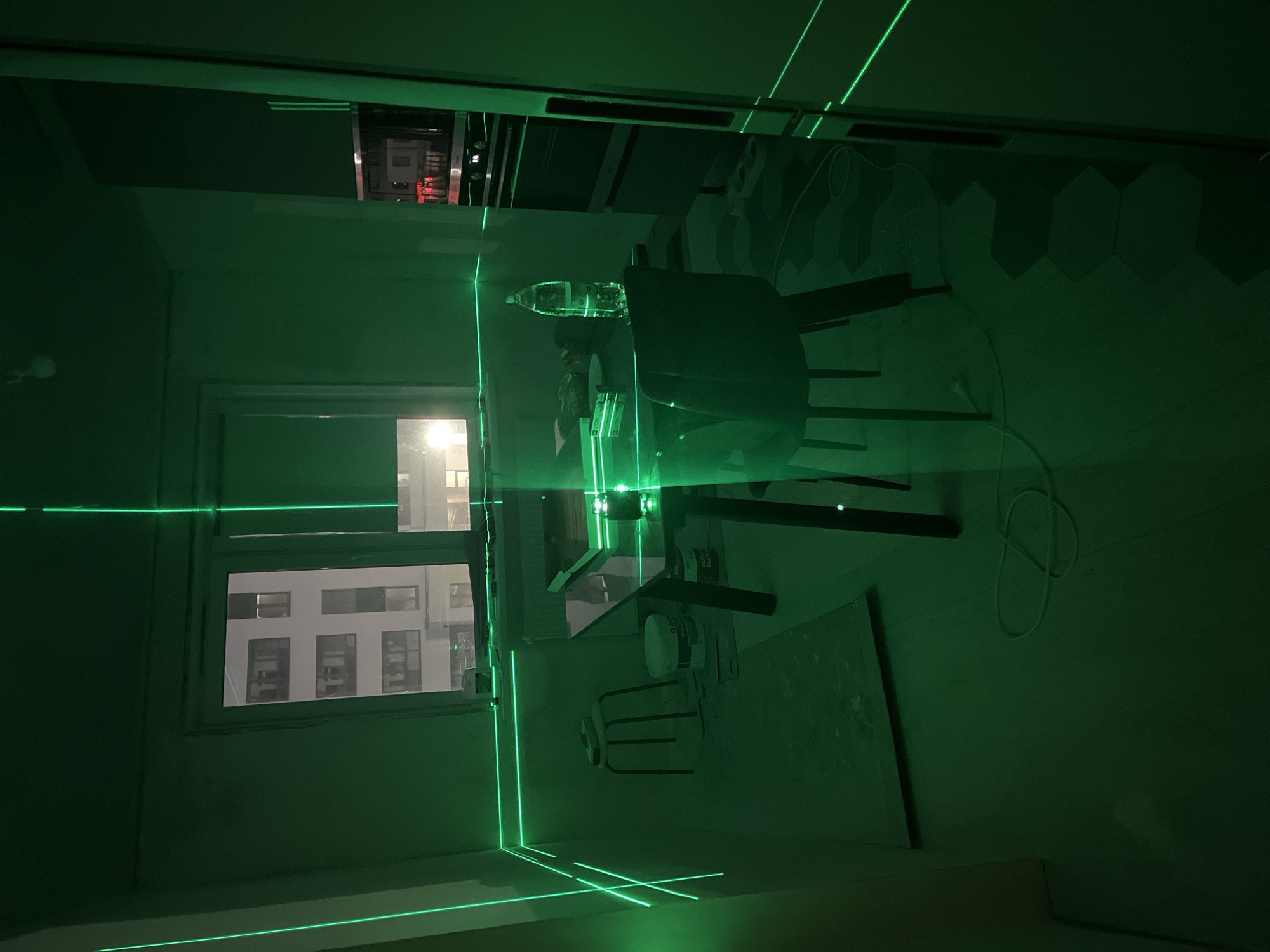 The image depicts an interior space, likely a room under construction or renovation, with green laser lines projected across various surfaces. The room contains a table with a water bottle on it, a chair, a window with a view of a multi-story building, and several paint cans. The room appears to be lit by the laser level, which creates a bright green pattern across the room and makes the lighting conditions quite dim. The city of Verkhnyaya Pyshma, Russia, cannot be confirmed or denied from the image.