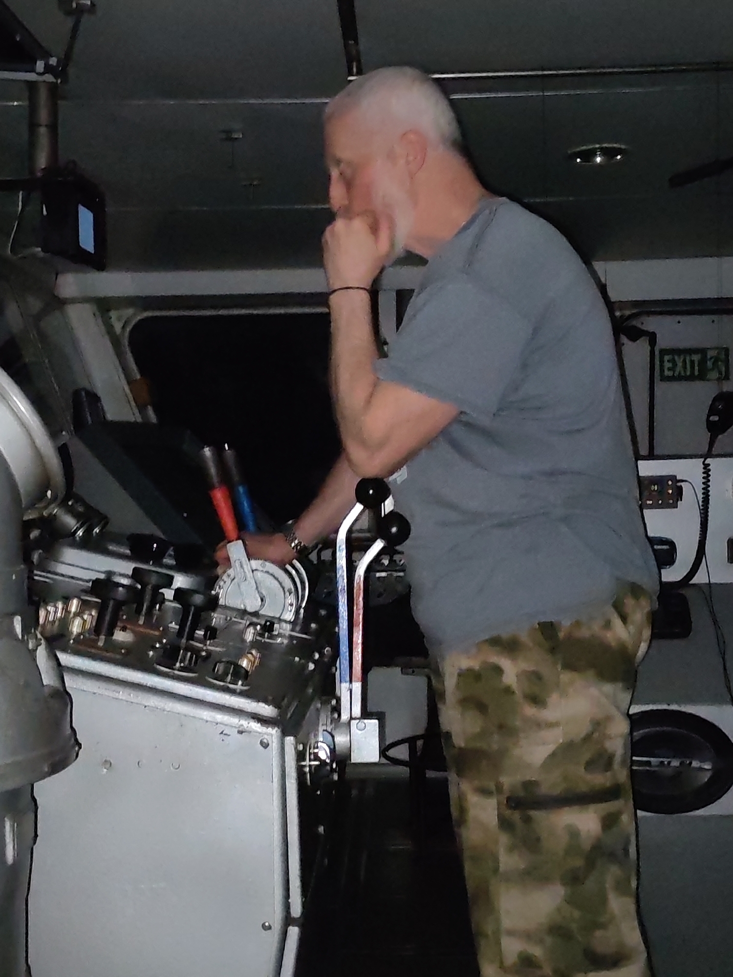 An adult male with white hair and a beard stands inside what appears to be the control room of a vessel. He is wearing a gray t-shirt and camouflage pants. His right hand is on his face, near his chin, while his left hand manipulates a lever on a large, metallic control panel. Various levers, switches, and gauges are visible on the panel. An "EXIT" sign is on the wall behind him. The location is Unknown, Unknown.