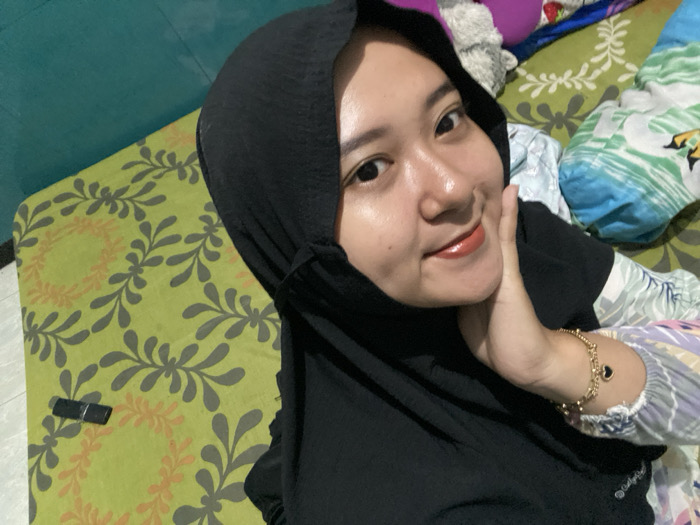 A young woman wearing a black hijab poses for a selfie. She's on a bed with a green, patterned sheet. Her hand rests gently against her cheek, displaying a gold bracelet with a heart charm. Her skin is clear and her lips are coated in a glossy, brownish-red lip color. A dark, rectangular object lies on the bed near her. The background includes pillows and blankets.