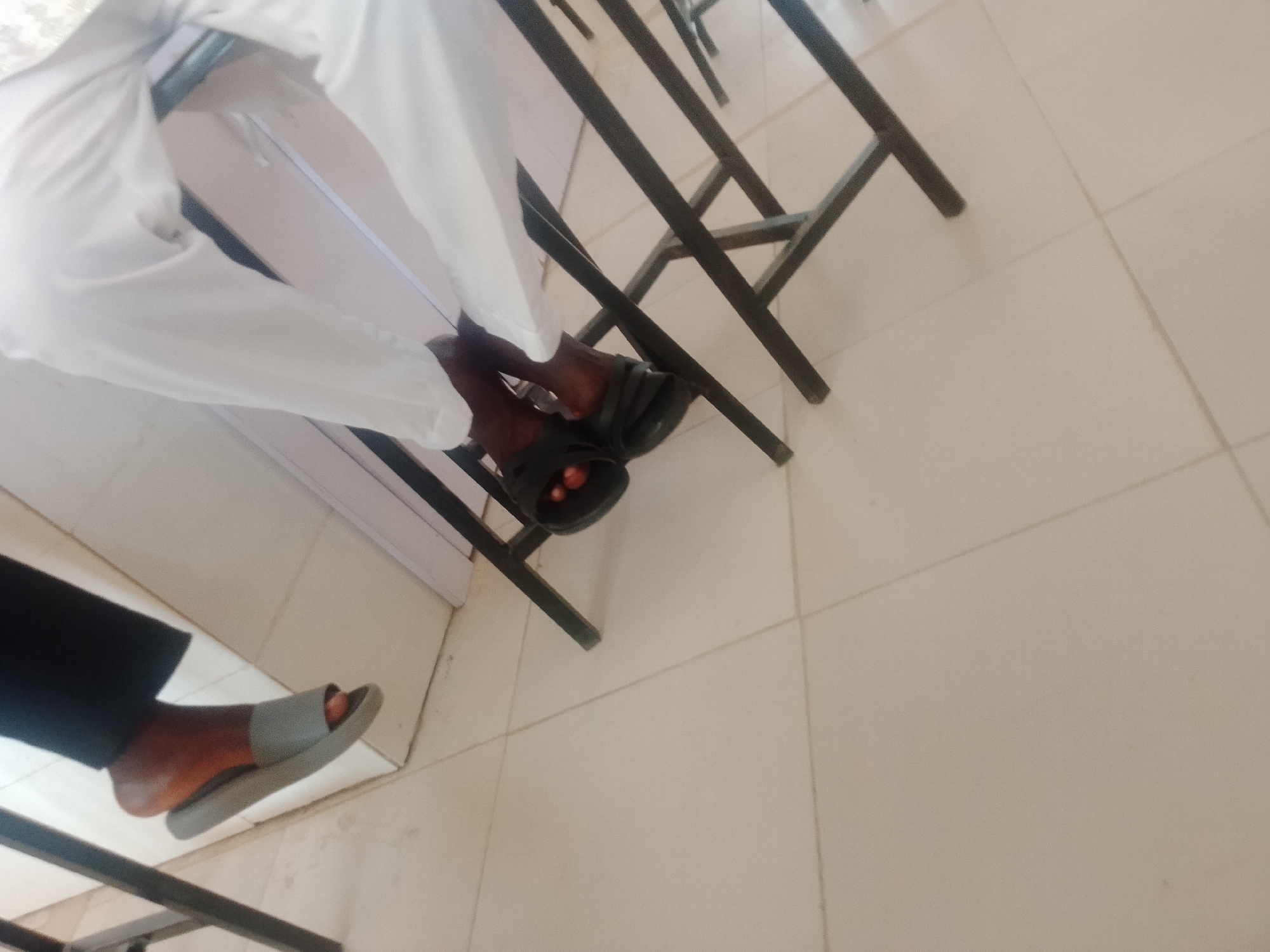 The image shows a person's legs and feet wearing white pants and dark sandals. The person is seated, with their legs crossed at the ankles. The scene appears to be indoors, with a tiled floor and a metal structure in the background. The lighting is somewhat dim, suggesting it might be an indoor space with artificial lighting.