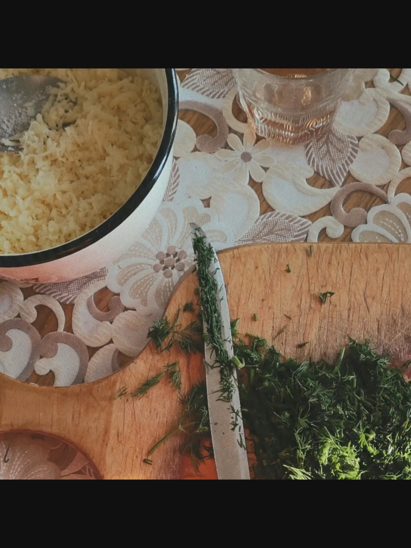 A close-up shot shows a bowl of finely grated cheese and a wooden cutting board with chopped dill and a knife. The bowl, with a black rim, is filled with light yellow grated cheese. A silver spoon is partially visible in the cheese. The cutting board, made of light-colored wood, has a pile of chopped green dill on its right side. A silver knife with a black handle, also partially visible, lies across the dill. In the background, a doily with a floral pattern in shades of cream and brown covers a wooden surface. A small glass containing a light amber liquid is placed on the doily. The lighting is soft, and the overall scene is well-lit, with a slight vignette effect. The image does not contain any information that would allow for identification of a specific location in New York City, United States of America.