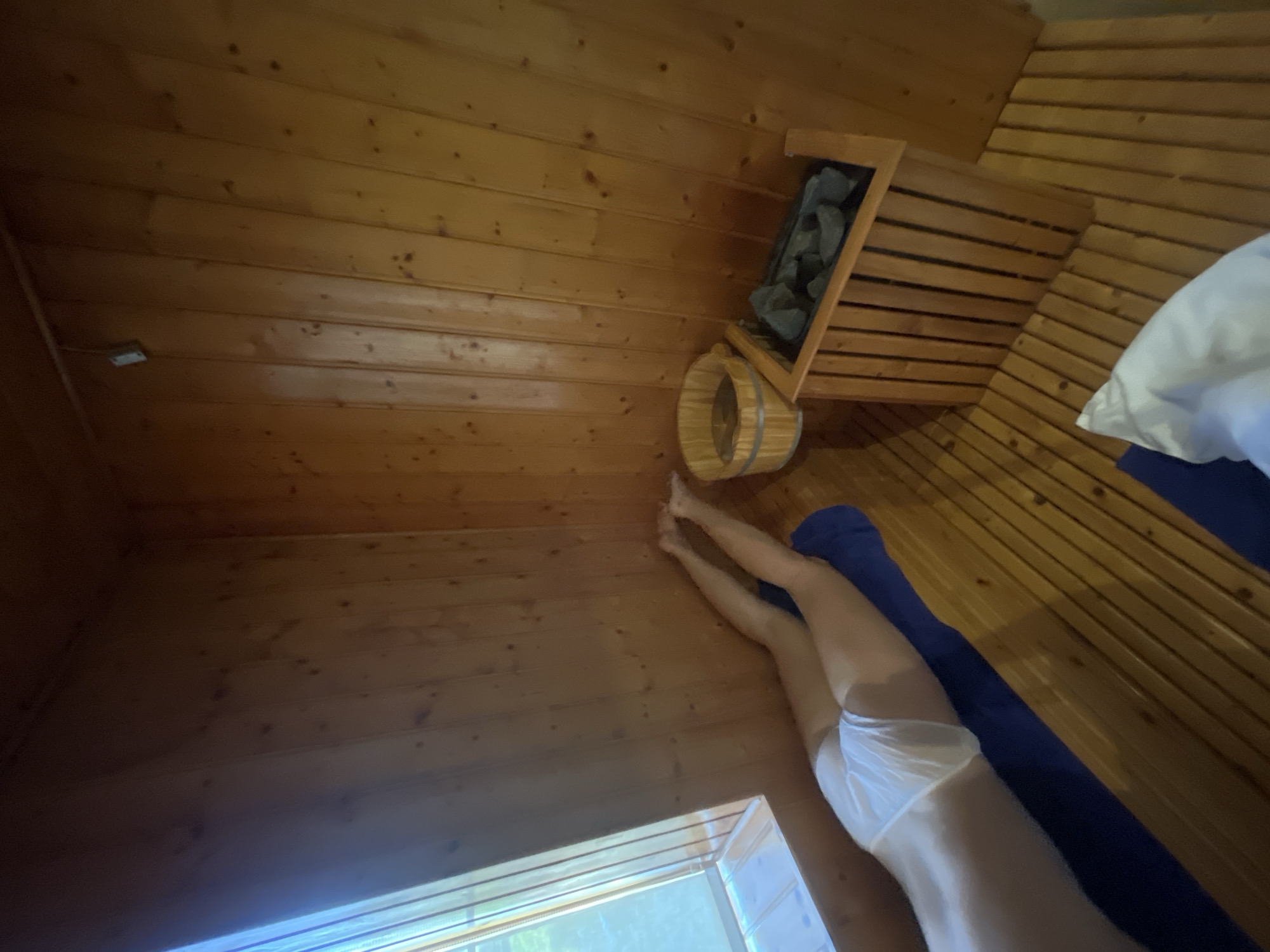 In a wood-paneled sauna, a person lies prone on a blue mat, wearing white underwear. Their legs are extended, and their feet are visible. To the right, a wooden sauna heater filled with dark stones stands on the slatted wooden floor. A wooden bucket rests near the heater. Sunlight streams in from a window on the left. The location is a sauna in Đà Nẵng, Vietnam.