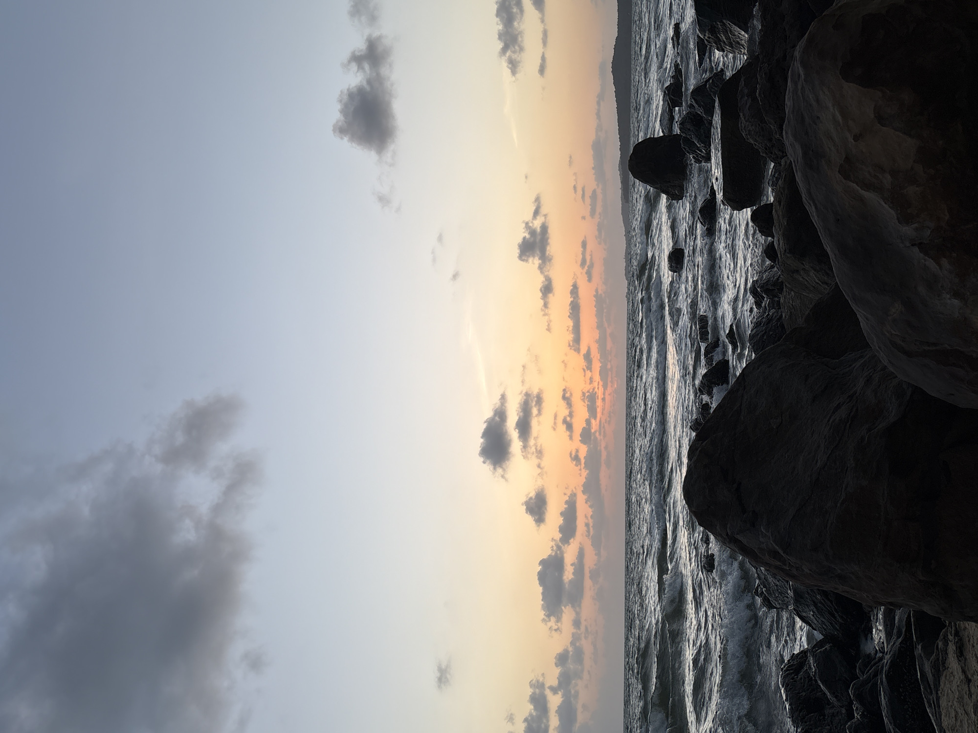 The image depicts a serene seascape with a rocky shoreline under a twilight sky. Large, dark rocks of various shapes and sizes are scattered across the foreground, some partially submerged in the water. The ocean exhibits gentle waves with white foam breaking against the shore. The sky transitions from a soft pastel gradient of orange and yellow near the horizon to a pale blue and grey higher up, with scattered clouds. In the distance, a faint outline of landmass is visible. The scene appears to be a natural coastal environment.