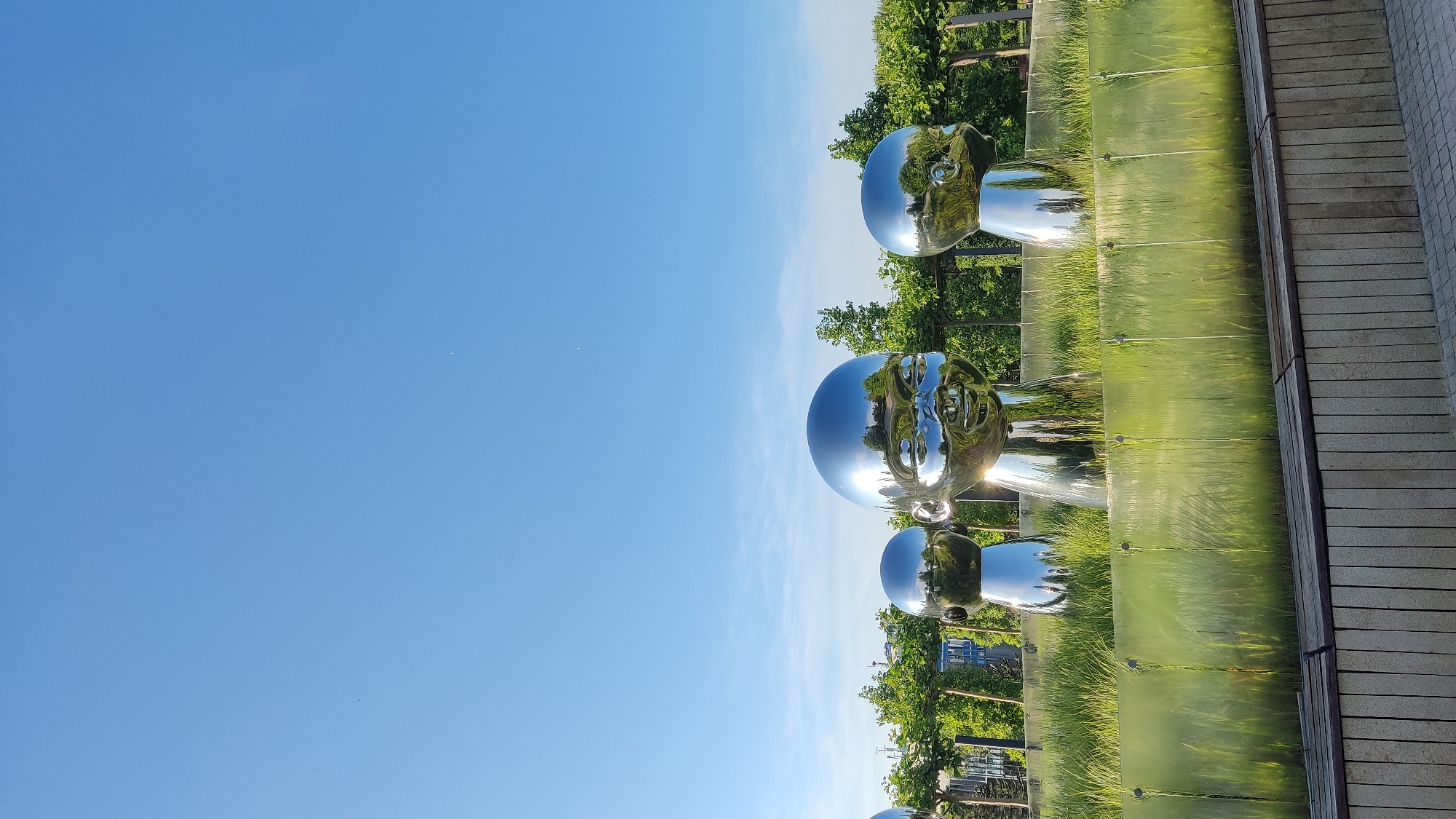 Three large, reflective, chrome-plated sculptures of baby heads are positioned in a grassy area with a clear blue sky above. The sculptures vary in size, with the central one being the largest and most detailed. They are positioned on a slightly raised bed of green grass, separated by a transparent pane of green-tinted material. In the background, a line of trees forms a backdrop, with some hints of buildings and structures visible beyond them. In the foreground, a wooden bench sits on a concrete base. The scene appears to be an outdoor public space. Based on the visual cues, this is the sculpture garden "Europe" in Krasnodar, Russia, featuring the "Baby" installation by artist Zhang Huan.