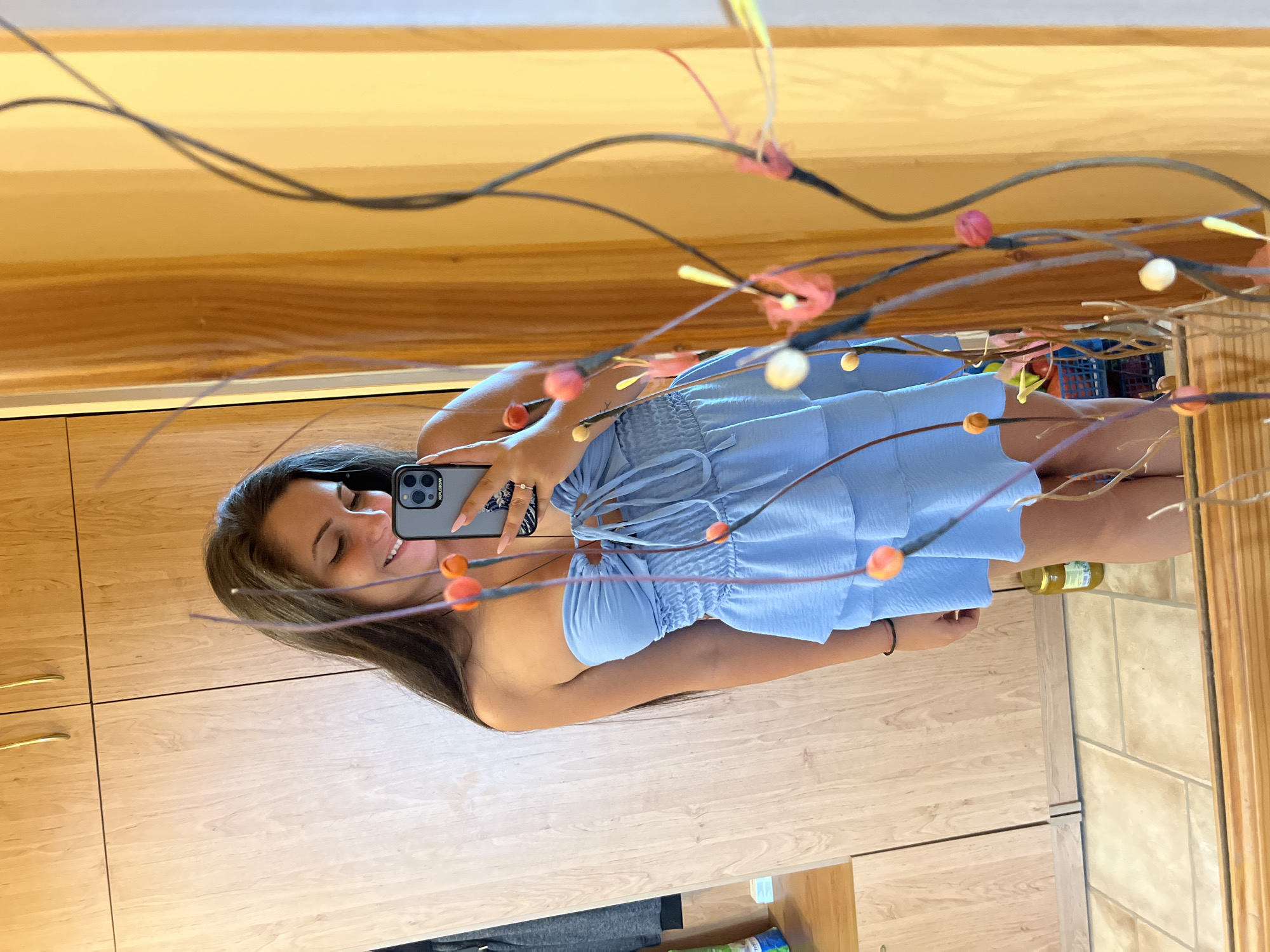 A young woman with long, dark hair is taking a mirror selfie in what appears to be a bedroom. She is wearing a light blue, strapless dress with ruffles. Her left hand is holding her smartphone, a silver iPhone with a black case, in front of her chest, angled to capture her reflection. Her right arm is by her side. She is smiling and looking at her phone's screen. The background includes a wooden wardrobe with gold handles, a mirror reflecting the room, and a decorative plant with small, colorful balls. The floor is tiled. The scene appears to be indoors, possibly in an apartment or house. There is no information in the image to suggest it is located in Vratsa, Bulgaria.