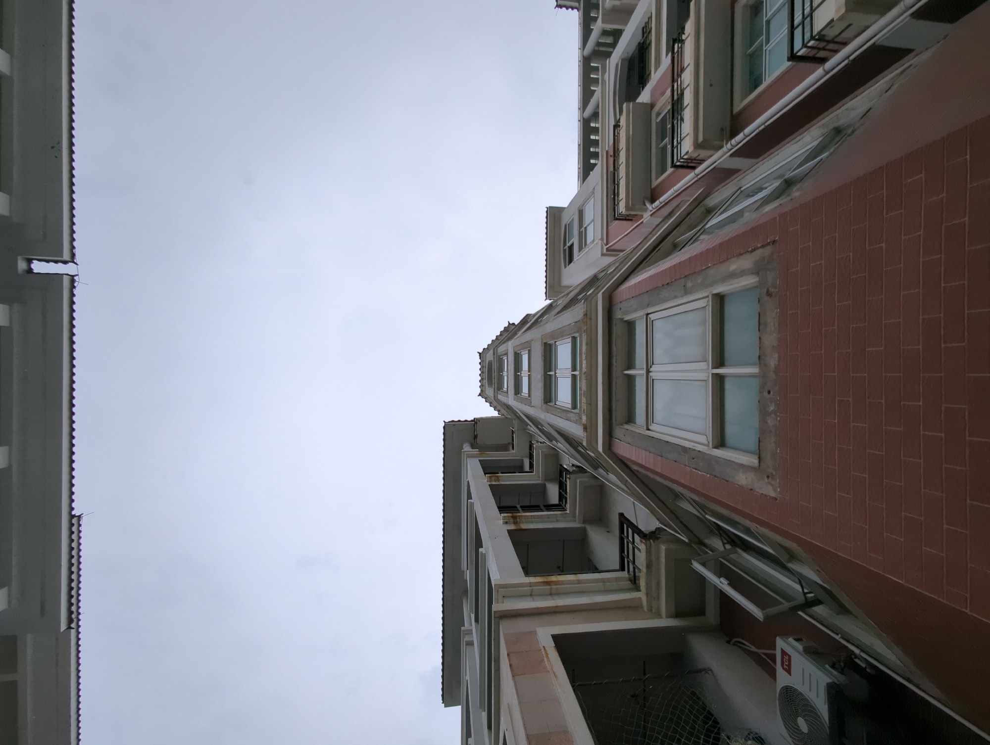 The image shows a low-angle shot of two buildings against a cloudy sky, likely in Shunde District, China. The building on the right is brick-faced with white-framed windows, some open. An air conditioning unit is visible on the lower right. The building on the left has balconies with what appears to be netting, supported by white columns. The perspective emphasizes the height of the buildings. No people are visible.