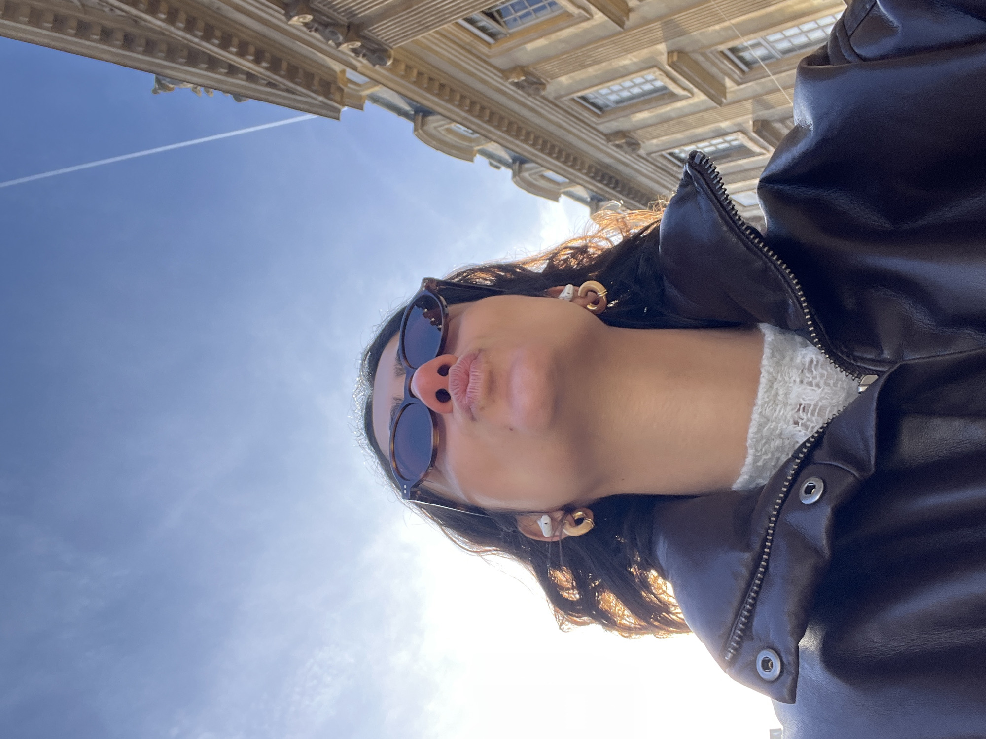 A woman wearing sunglasses and a brown leather jacket poses for a selfie in Paris, France. The camera is angled upwards, showing the woman with a building and a blue sky in the background. A contrail is visible in the sky. The woman has dark curly hair, wears earrings, and has her lips pursed.