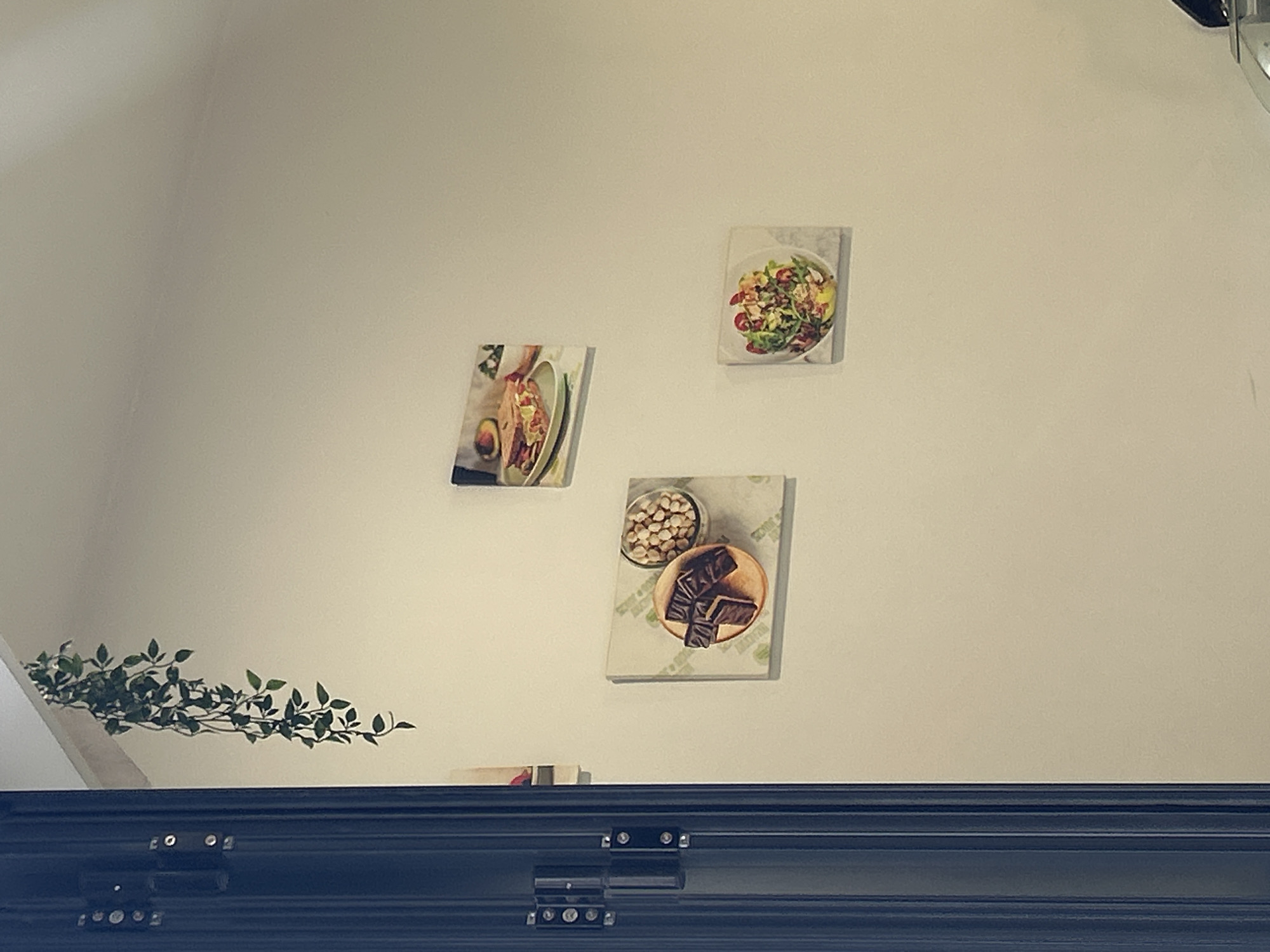 The image shows a white wall with three food-related pictures hanging on it. From top to bottom, there is a salad, a sandwich, and a plate of chocolate and nuts. There is a green plant in the bottom left corner and a black molding across the bottom of the image. The location is in a building, in a city of Unknown, Unknown.