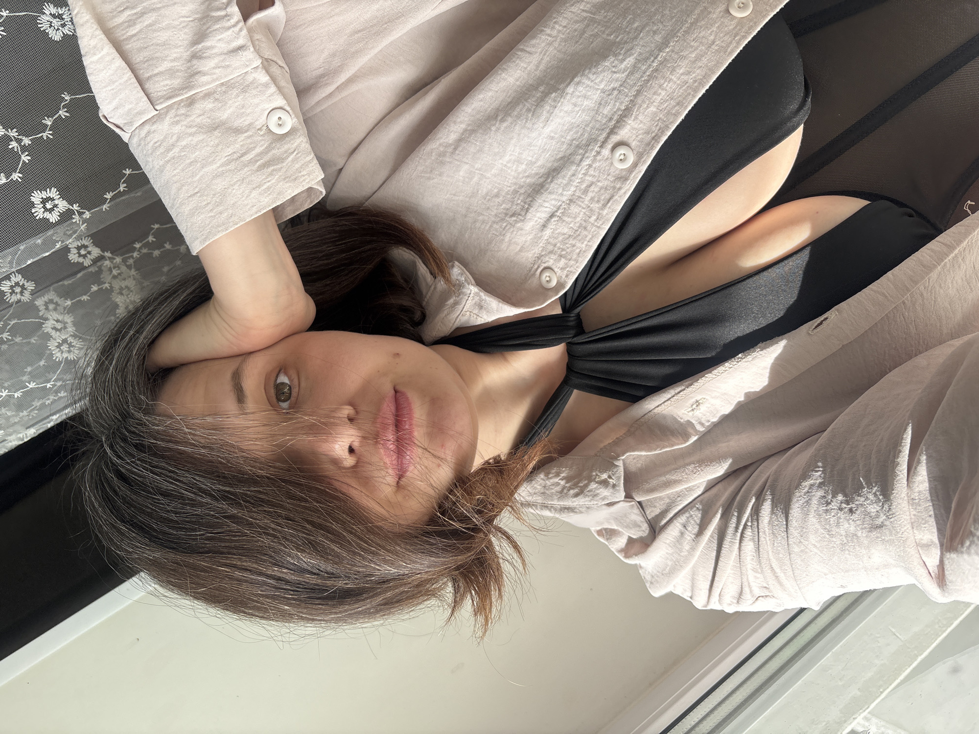 A woman with shoulder-length brown hair lies on her side, her head resting on her hand. She wears a light-colored, button-down shirt over a black garment, possibly a bathing suit top. The scene is illuminated by sunlight, casting shadows on the woman's face and clothing, and on the window frame. The location is inferred to be Chaikovsky, Russia.
