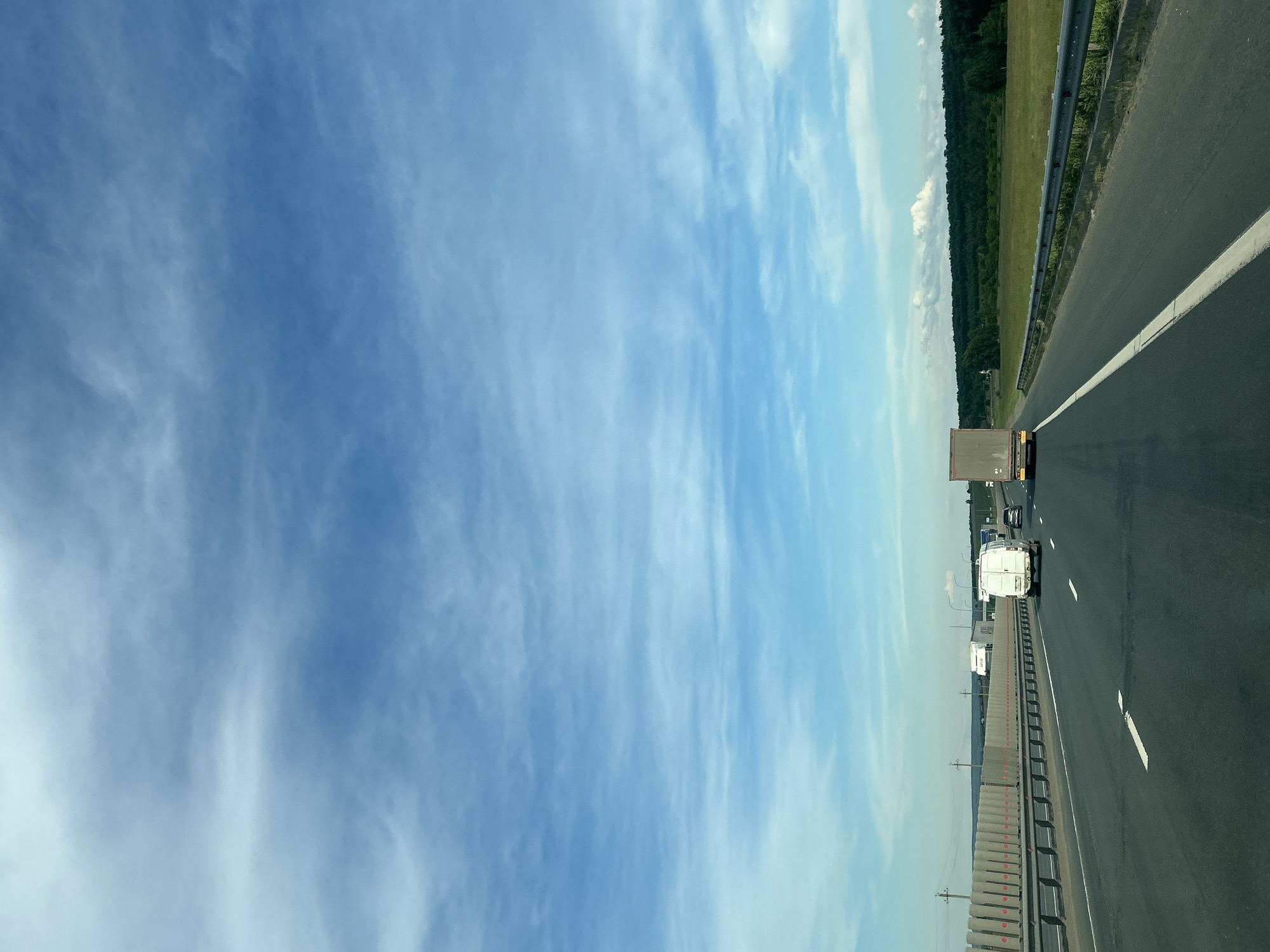 The image depicts a highway in Malaya Tsarevschina, Russia. Several vehicles, including trucks and a van, are traveling on the roadway. The sky is a light blue with wispy clouds. To the right of the highway is a green field and a treeline. The road has a concrete barrier on the left and a guardrail on the right. White lane markings are visible on the road surface.