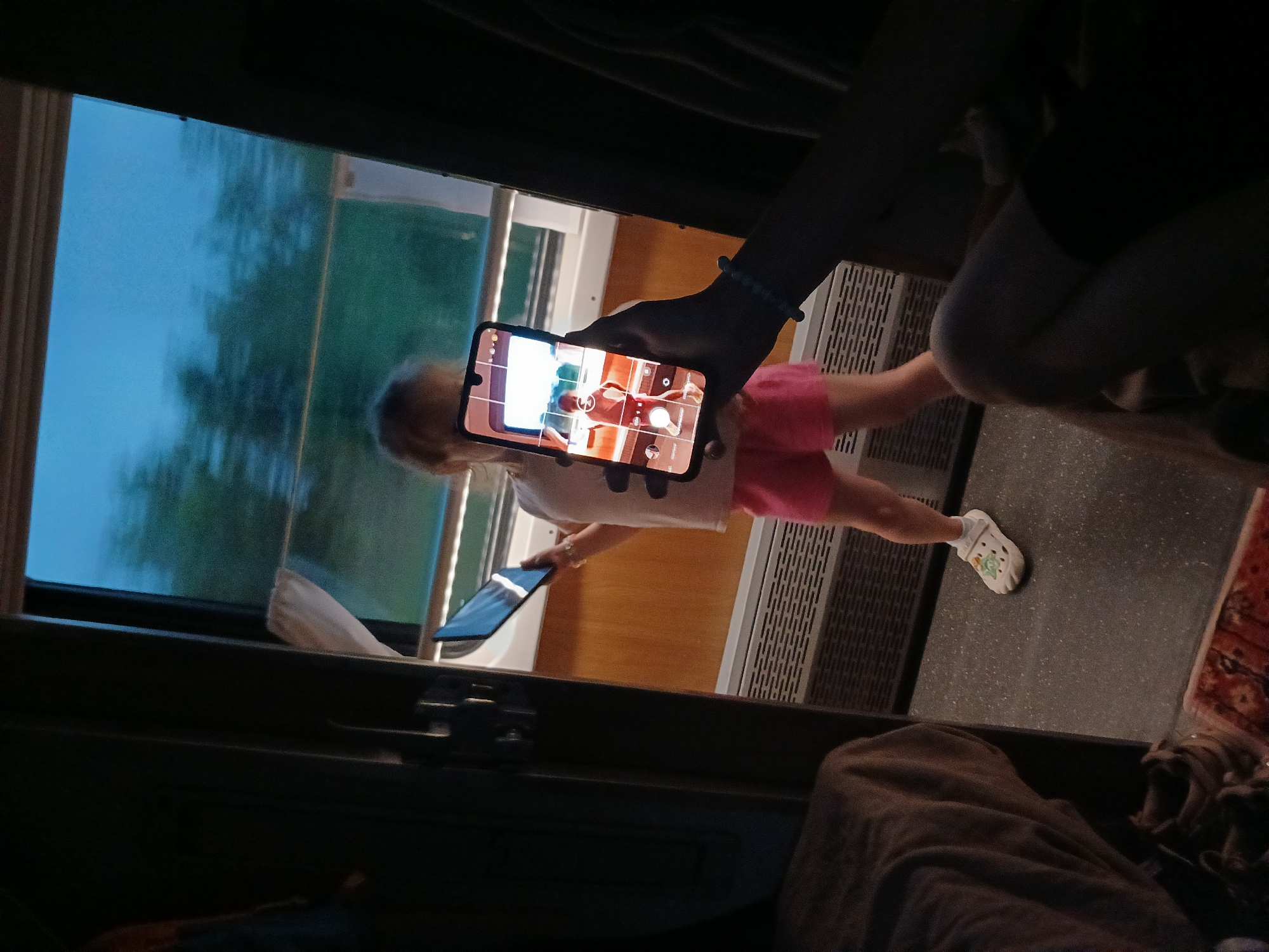 In a dimly lit train compartment, a young girl in pink shorts and a white t-shirt stands by the window, holding a tablet. Her back is to the viewer, and she appears to be looking out at the passing scenery. In the foreground, a hand holds a smartphone, its screen displaying a live camera feed of the girl. The phone's screen shows the girl in a red top and pink shorts, with a blurred background suggesting motion. The compartment's interior is visible, with luggage draped over a seat and a rug on the floor. The window shows a hazy, blueish sky and green trees, indicating that the train is in motion, possibly traveling through the countryside near NovoYa kupovo, Russia.
