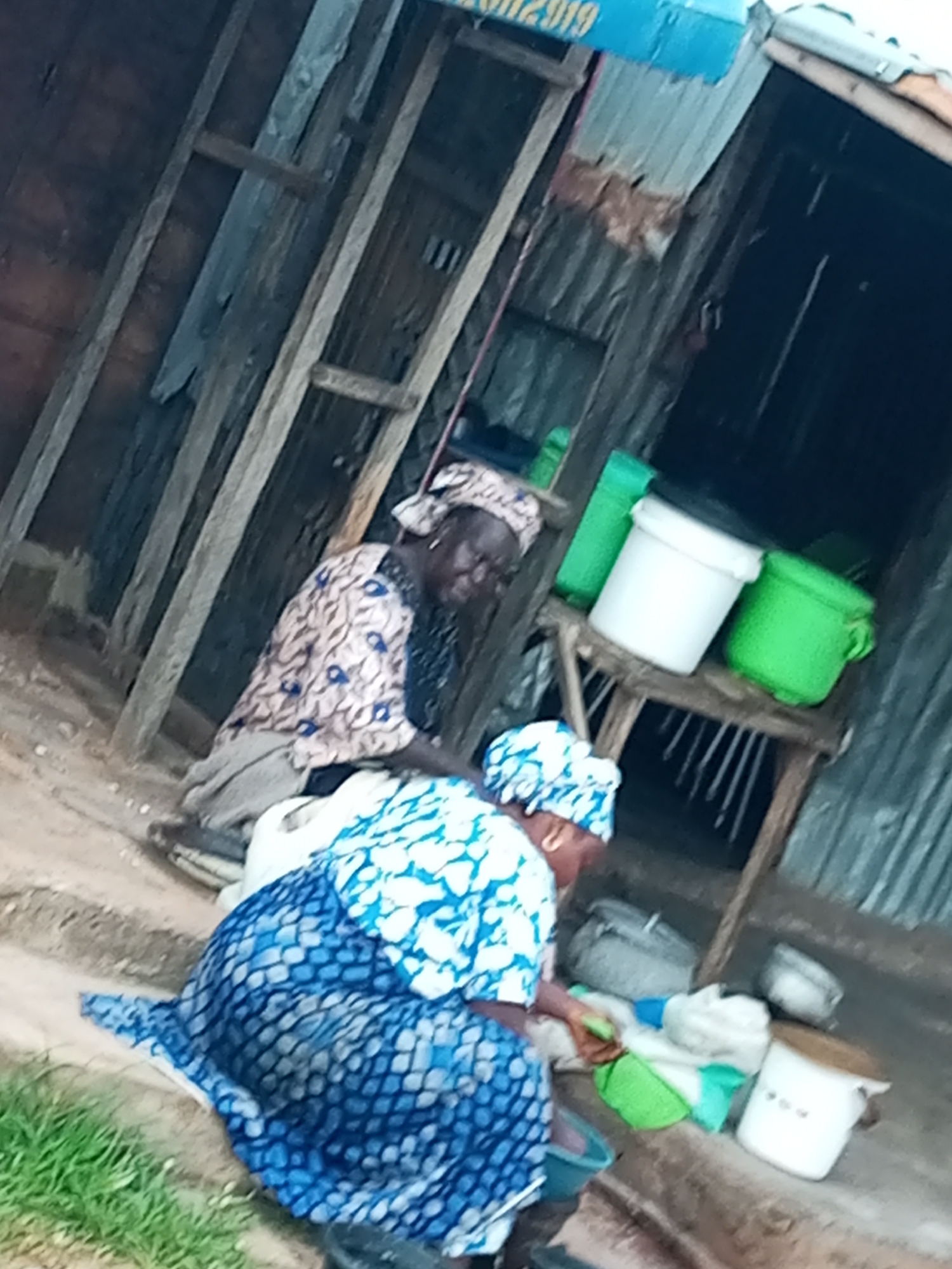 Two individuals are engaged in domestic chores near a rustic, open-air structure in Tumfure, Nigeria. The structure appears to be constructed from corrugated metal and salvaged wood. In the foreground, a person wearing a blue and white patterned dress and head covering is kneeling and appears to be washing or sorting items on the ground, with a green basin to their left. Behind them, another person, also in traditional attire with a head covering, sits near a wooden platform holding several containers, including a white bucket and a green bucket. The ground is dirt, with some patches of grass visible. The overall scene suggests a rural or semi-urban domestic setting.