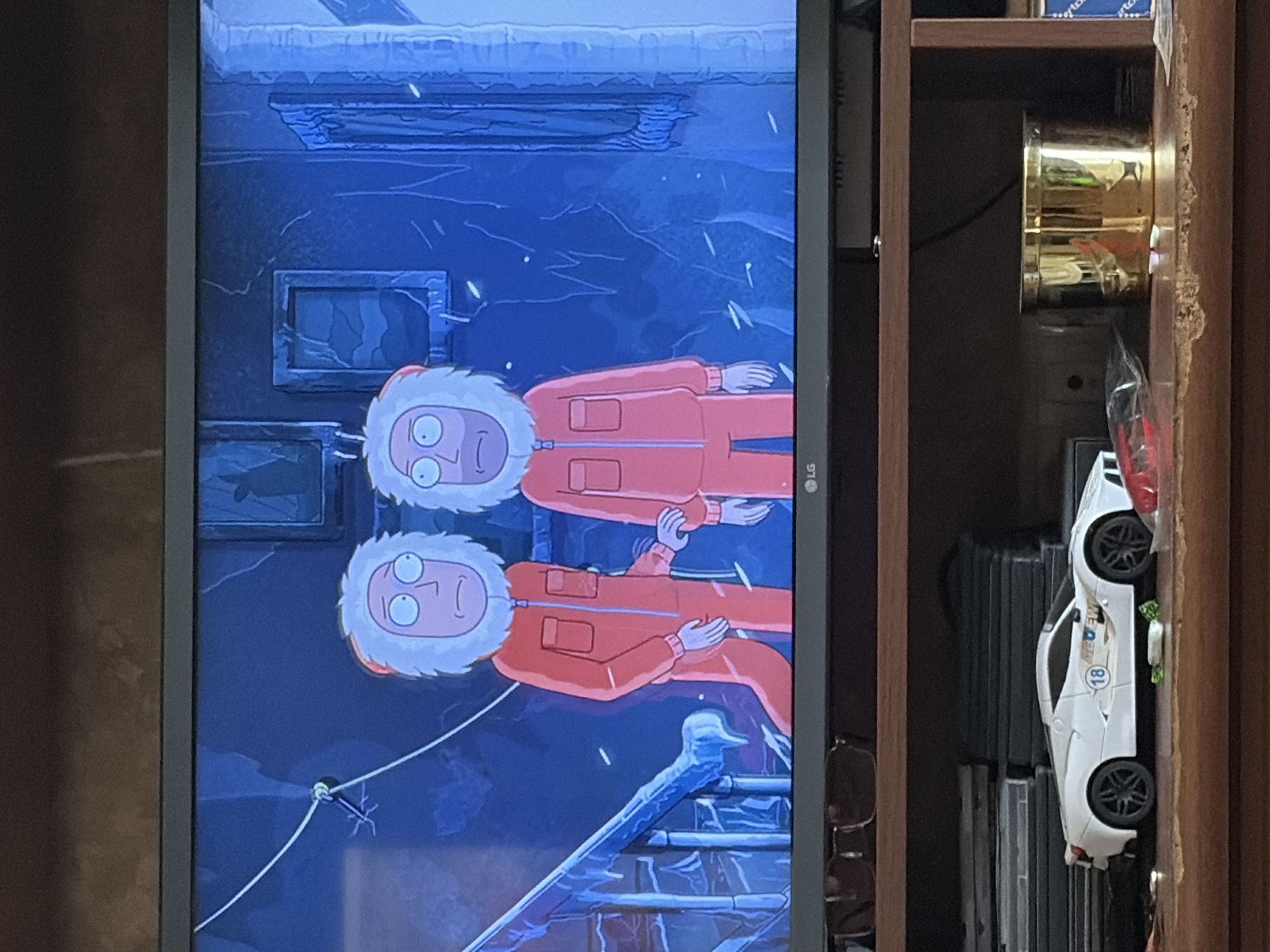 On a television screen, two animated characters resembling Rick Sanchez and Morty Smith are depicted in an icy environment. They are wearing orange jumpsuits with furry hoods and appear to be walking down a frozen staircase. The background shows icy walls and framed pictures. Below the television, on a wooden shelf, a white toy car and a golden canister are visible.