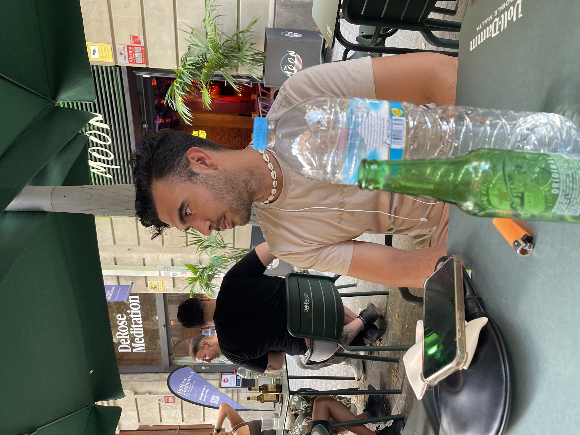 A medium shot captured outdoors in Barcelona, Spain, shows a group of people seated at a cafe table. The foreground features a mobile phone on a black bag, an orange lighter, a green glass bottle, and a clear plastic water bottle with a blue cap. In the midground, a man with dark hair and a light tan t-shirt wearing a seashell necklace is looking to his left. Behind him, a man in a black t-shirt is seated. The background displays the facade of a building with signs for "DeRose Meditation" and "MOON." Green umbrellas and potted plants are visible, contributing to the outdoor cafe atmosphere.