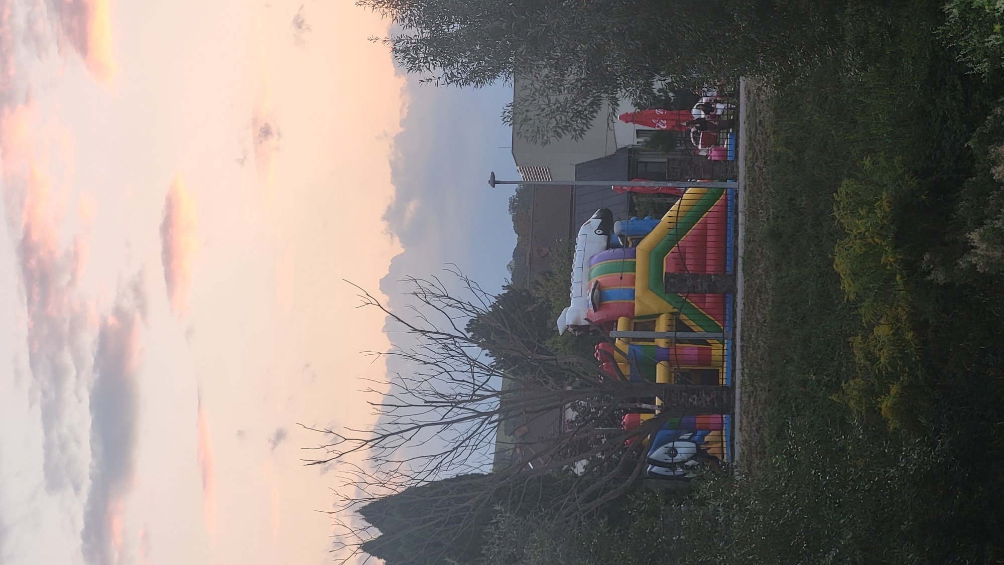 In the city of Prešov, Slovakia, an inflatable playground shaped like a rocket ship is visible behind a fence. The playground is brightly colored with red, yellow, green, blue, and purple sections. A bare tree with many branches is in the foreground to the left of the playground. Behind the playground, buildings and trees are visible, and the sky is filled with pastel-colored clouds.