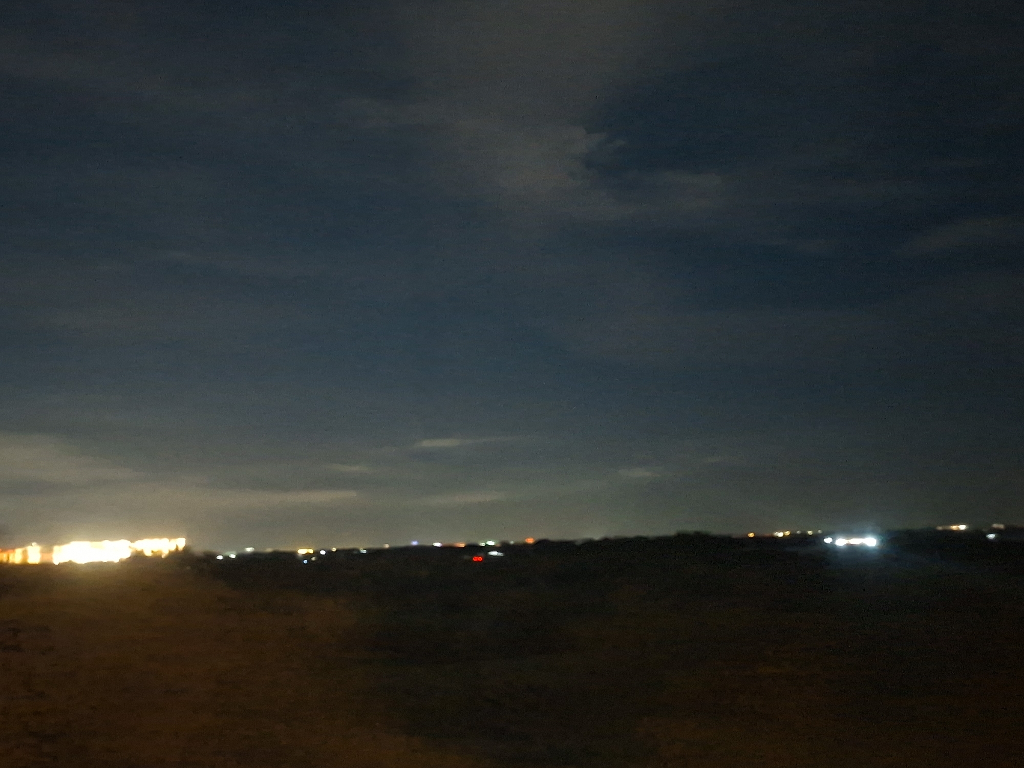 The image captures a nighttime scene with a dark, cloudy sky. In the distance, a line of lights suggests a populated area, possibly a city or town. The lights are blurry and appear to be from buildings and vehicles. The foreground is dark and appears to be unpaved ground. The overall impression is one of a rural or semi-urban landscape at night. The location is identified as Thủ Đức, Vietnam.