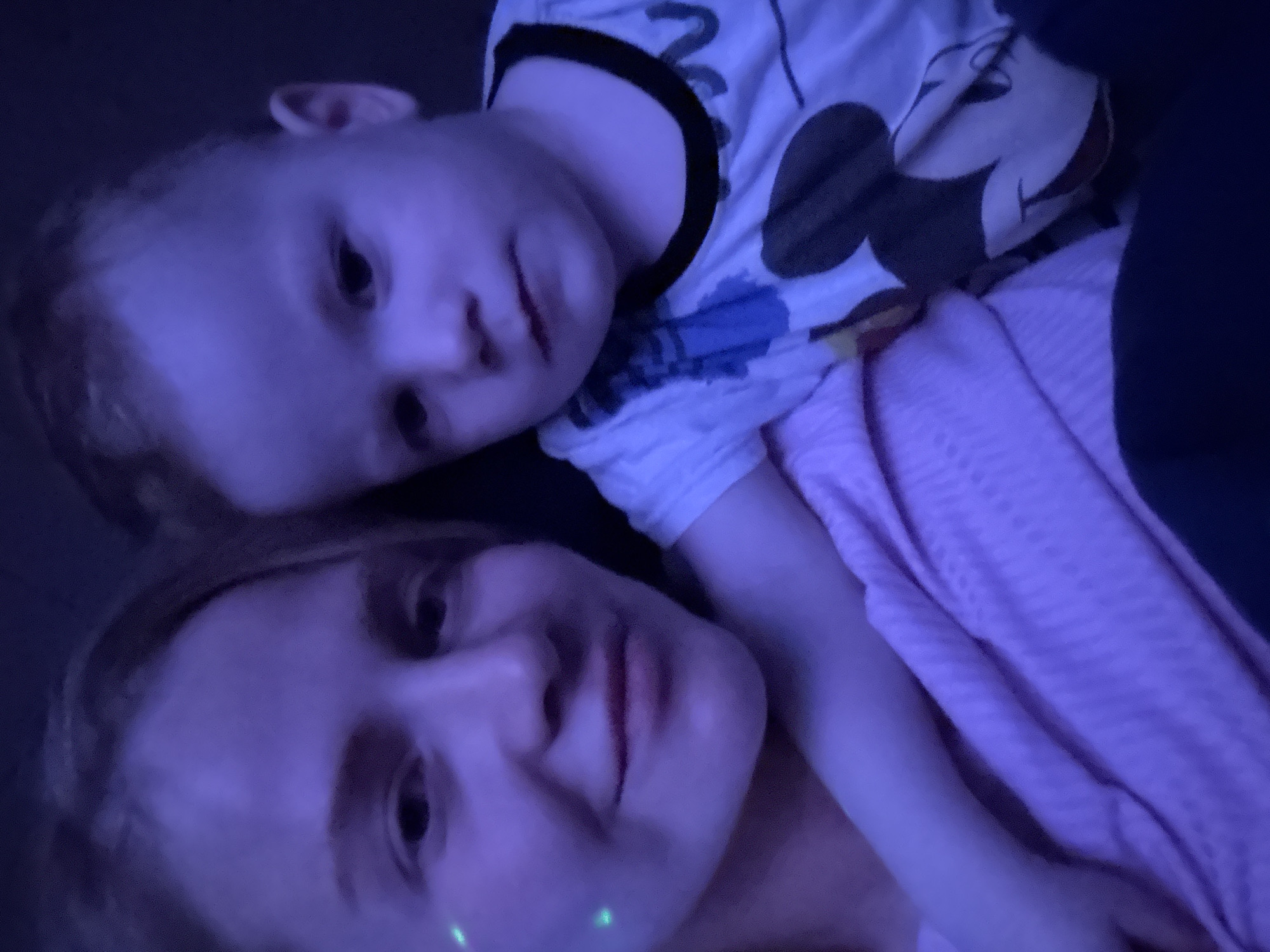 A woman and a young child lie together. The woman is on her back with the child resting on her chest. The child is wearing a white shirt with a black collar and a Mickey Mouse design. The woman is wearing a pink shirt. There is a green light visible on the woman's face. The lighting in the image is predominantly blue. The location is not discernible from the image.