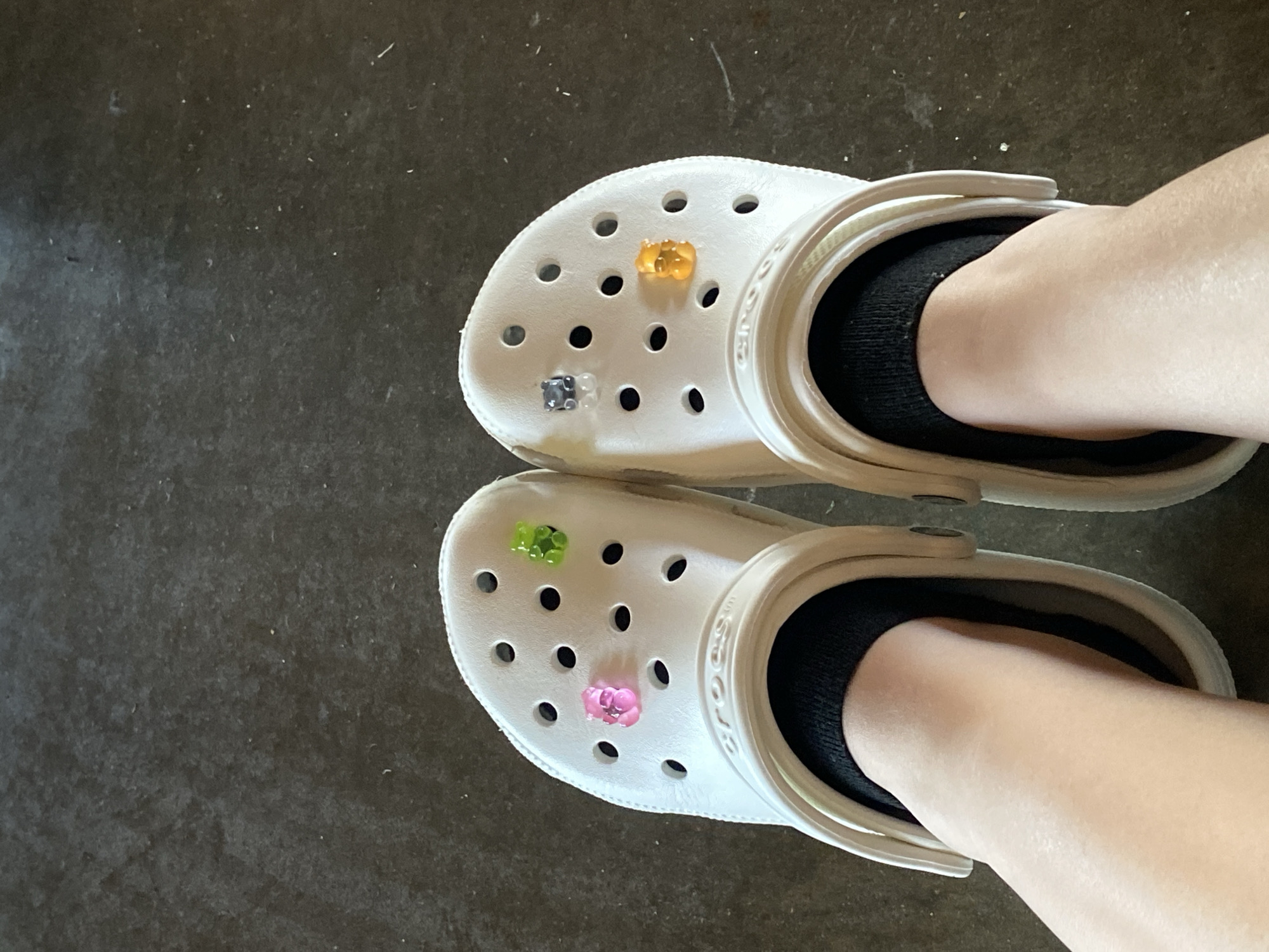 A pair of white Crocs adorned with gummy bear-shaped charms are shown from a top-down perspective, worn by a person with black socks on their feet. The Crocs are decorated with a pink gummy bear on the left shoe and a green, gray, and orange gummy bear on the right shoe. The shoes rest on a dark, textured surface, possibly a floor. The image appears to be taken indoors, in Washington Township, United States of America.