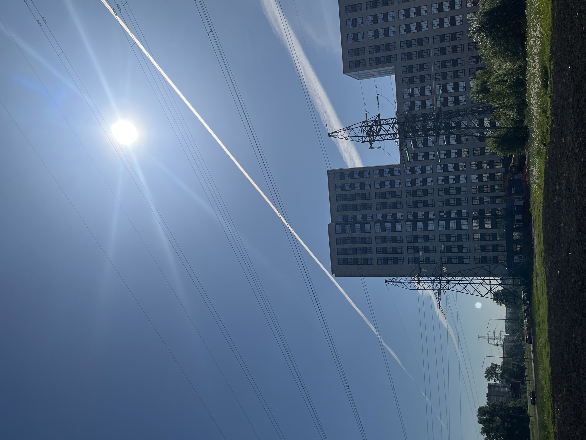 This portrait-oriented image captures a bright, sunny urban or suburban landscape in Saint Petersburg, Russia. The scene is dominated by a brilliant blue sky, across which numerous thin power lines stretch, connecting to large, metal lattice power pylons. A dazzling sun, emitting strong rays, is positioned in the upper left, while two prominent white contrails streak across the clear expanse.
On the right side of the frame, two tall, modern multi-story buildings, likely residential or office, with numerous windows, rise towards the sky. A large power pylon stands prominently between these buildings in the mid-ground, with another visible further in the distance. Scattered green trees and vegetation are seen behind and around the structures, and a patch of green grass occupies the immediate foreground on the right. Towards the lower left, a road with a few distant cars and streetlights is faintly visible, and a yellow excavator is partially discernible near the base of one of the pylons.
The image depicts a clear and bright daytime, suggesting either mid-morning or afternoon. No specific human activity or event is taking place, presenting a static snapshot of urban infrastructure and nature under a strong sun. No visible text is present in the image.