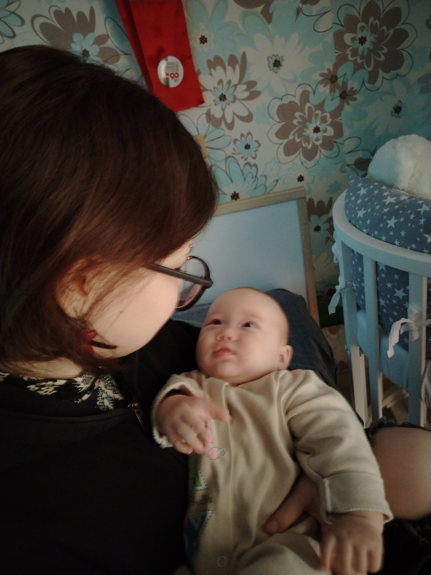 A woman with brown hair and glasses holds a baby wearing a beige onesie. The woman is looking down at the baby. The background is a floral wallpaper and a white crib with a blue star patterned cushion. There is a red object with a white circle and number on it hanging on the wall. The location is potentially in a home within the city of Гожан, Russia, but this information is not directly discernible from the image itself.