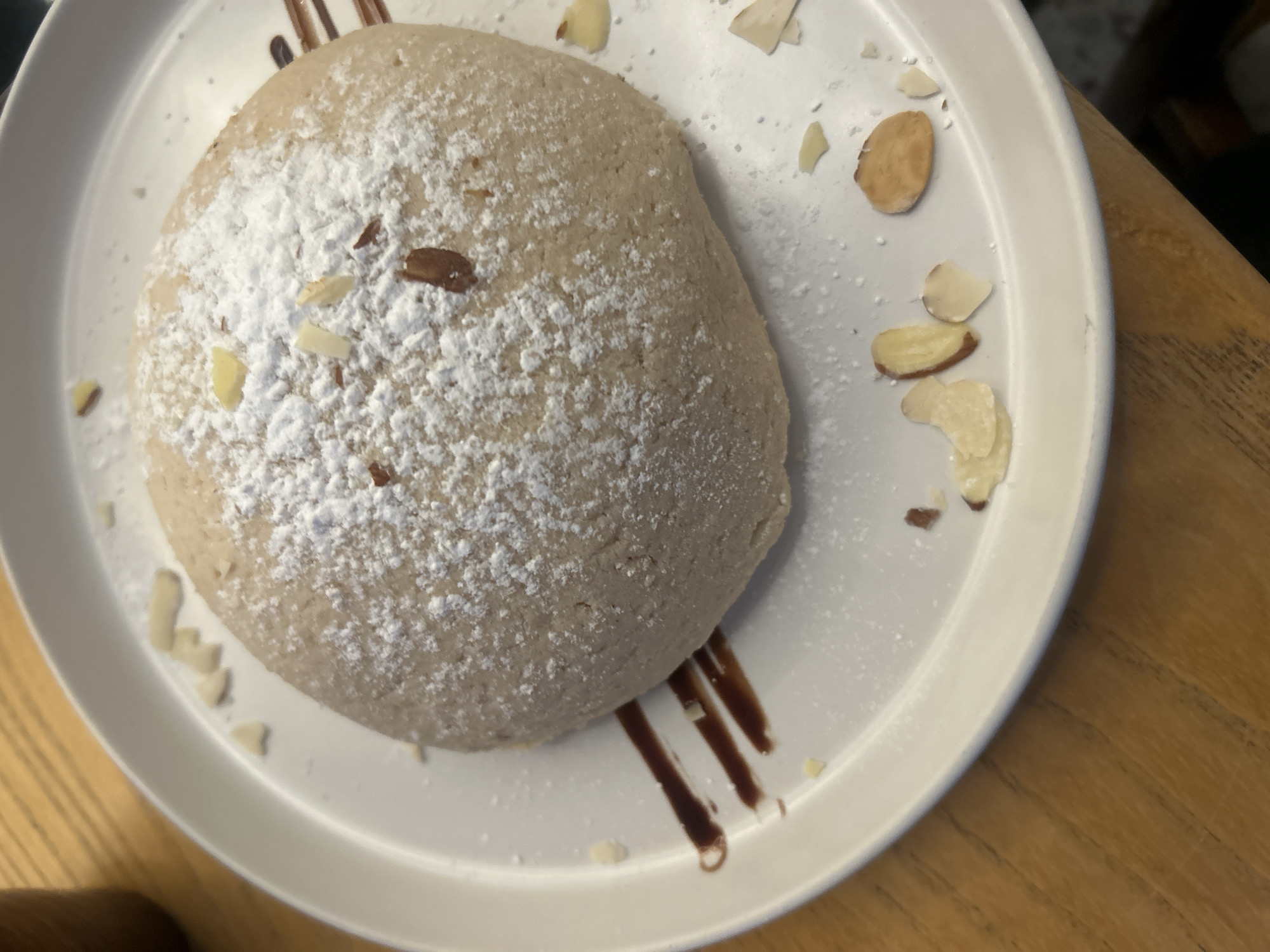 A white plate is viewed from above, containing a tan-colored, dome-shaped cake dusted with powdered sugar. The cake is garnished with scattered sliced almonds. There are drips of dark chocolate sauce on the plate. The plate rests on a wooden surface. The location is Unknown, Unknown.