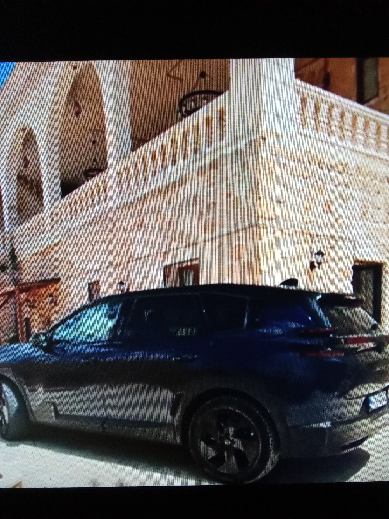 The image shows a dark blue SUV parked in front of a beige building with arched windows and a balustrade. The building appears to be made of stone or stucco and has a textured surface. There are decorative elements on the building, including what look like outdoor light fixtures and a wrought-iron railing. The car is seen from the side and slightly from the rear, with its dark wheels and tinted windows visible. The ground in front of the car is paved. The overall impression is of a vehicle parked in an outdoor setting with traditional architecture. The location is specified as Рощин ауылдық округі, Kazakhstan, however, the visual elements do not provide specific clues to confirm or deny this location.