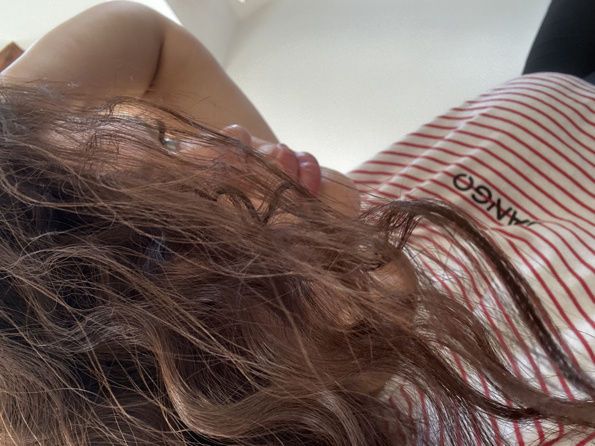 A close-up, low-angle shot shows the face and upper body of a young woman indoors. Her long, brown, wavy hair is partially obscuring her face, with strands falling across her eyes and forehead. Her eyes are looking to the left, and her lips are slightly parted, revealing a hint of her teeth. She is wearing a red and white striped t-shirt. The background is blurred and consists of a light-colored wall and what appears to be furniture. The image appears to be taken in a private residence in Katowice, Poland.