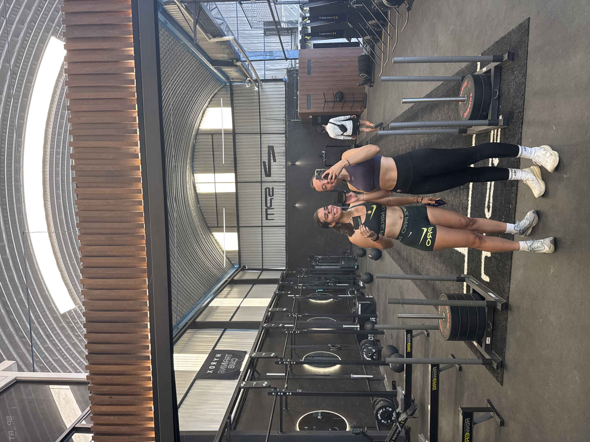 The image shows two young women in athletic wear taking a selfie inside a gym. They are standing side-by-side, facing the camera, with one holding a phone. The gym is equipped with various exercise machines and weights. A man is visible walking in the background. Banners with the text "HABOX" and what appears to be mirrored text are hanging in the background. The location is likely Barrio de Nuevo León, Mexico based on the text "MIKEGGO V" and other text at the bottom of the image.