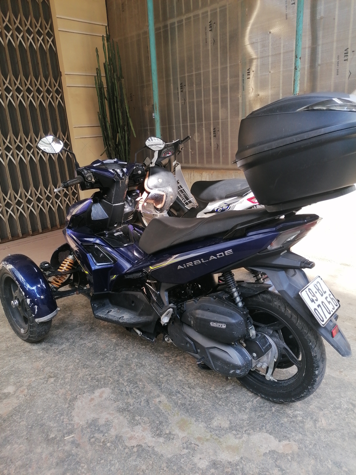 In Xuân Thành, Vietnam, a three-wheeled scooter with a dark blue body and a black cargo box on the rear is parked on a concrete surface. The scooter features a yellow suspension system on its front axle, and the brand name "AIRBLADE" is visible on its side. A license plate with the characters "49-BZ 070.56" is attached to the rear. To the left of the scooter, a collapsible metal gate is visible, and to the right, a portion of another scooter with a white and red body is partially obscured. Behind the scooters, a tall, green cactus stands next to a wall with a beige finish. The background also shows a transparent corrugated structure with vertical metal supports.