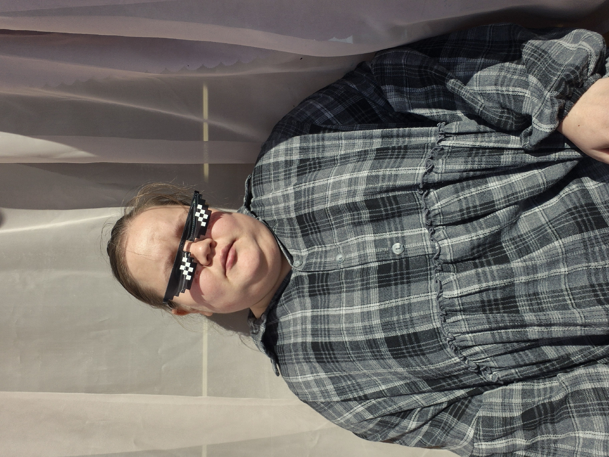 A medium shot features a fair-skinned individual wearing pixelated sunglasses and a gray and black plaid dress with ruffled detailing. The person is positioned against a backdrop of light-colored, semi-transparent fabric. The setting appears to be indoors due to the controlled lighting. No other individuals or significant objects are present. The exact location is not discernible from the image. The city of Chaikovsky, Russia, cannot be confirmed as the location based on visual evidence.