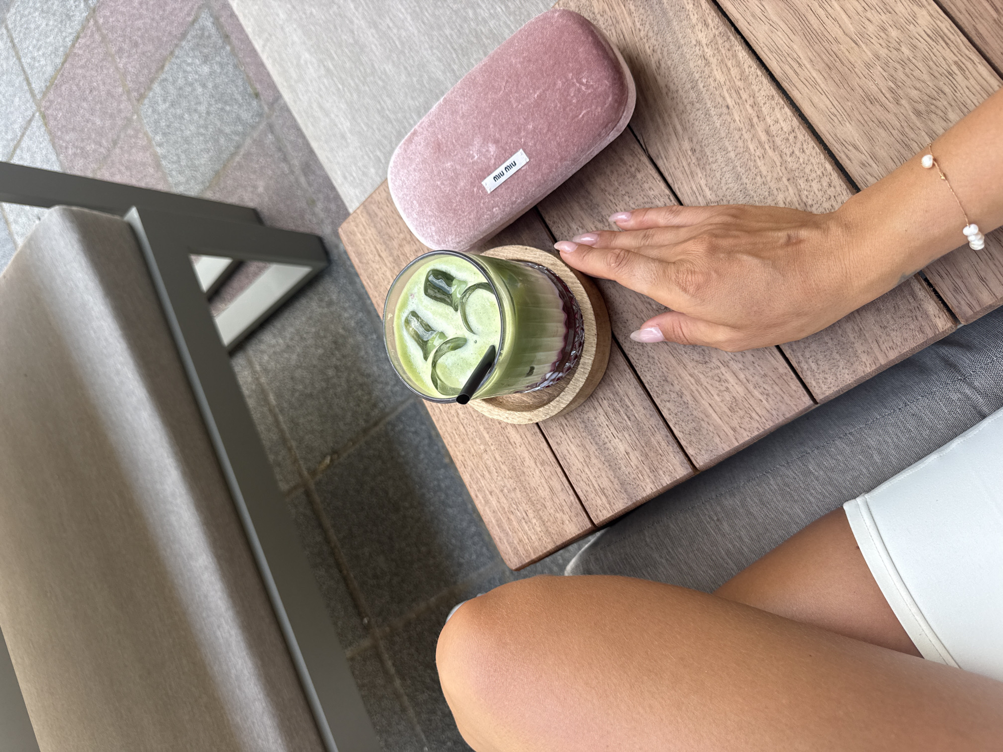 A person's bare leg and arm are visible in the foreground, resting on a textured surface that appears to be a patio or outdoor seating area. On a wooden table in front of them, there is a glass of green matcha tea with ice, a straw, and a small wooden coaster. To the right of the drink is a pink velvet glasses case with a small white Miu Miu tag. The background shows an outdoor seating area with a modern chair. The scene is set in the city of Fethiye, Turkey.