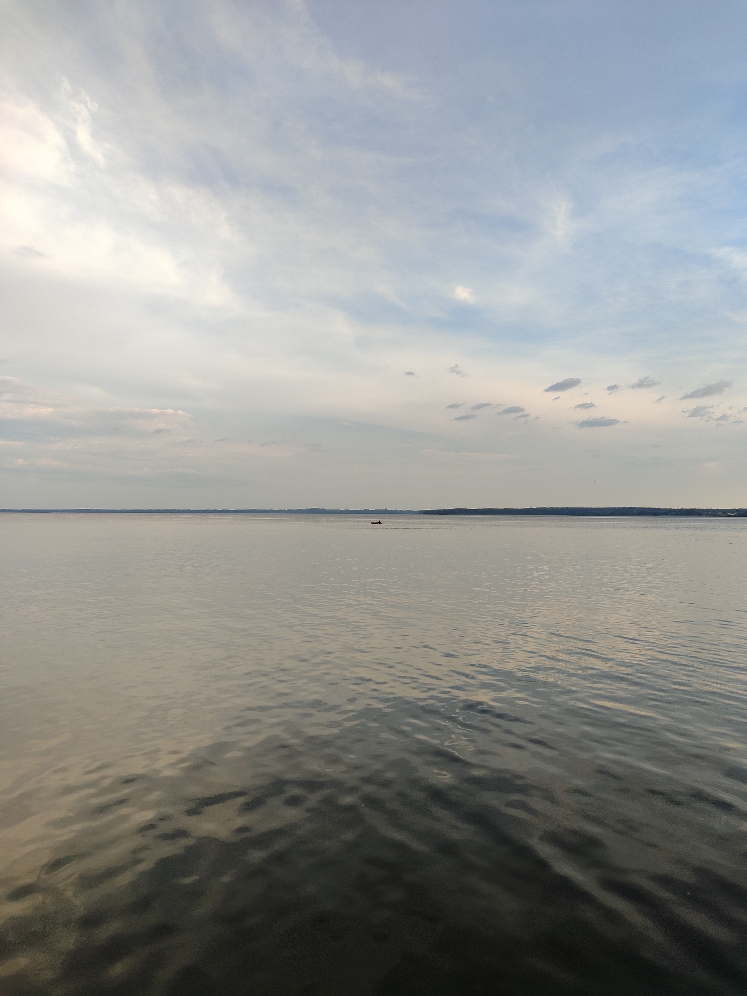 The image is a landscape photograph of a large body of water with a pale blue sky overhead, characterized by scattered clouds. The water's surface is calm, with gentle ripples reflecting the muted light from the sky. A thin, dark line of land is visible in the distance, suggesting a shoreline or the edge of a continent. On the water, a small, dark object, possibly a boat or a raft, is positioned slightly to the right of the frame's center, in the mid-distance. The overall atmosphere is serene and expansive.