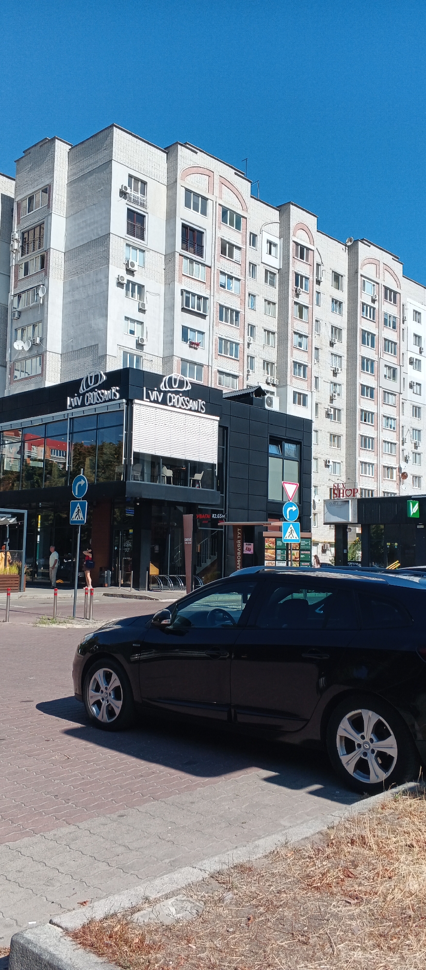 The image depicts a street scene in Cherkasy, Ukraine, dominated by a large, multi-story residential building with a light-colored brick facade and numerous windows. In the foreground, a dark-colored car is parked on a paved street. Adjacent to the street, a modern, black-fronted commercial building with large glass windows features signage that reads "LVIV CROISSANTS." Several traffic signs are visible, including a pedestrian crossing sign and a roundabout sign. People are visible on the sidewalk in front of the commercial building, and there is dry grass and weeds along the edge of the street and sidewalk. The sky is clear and blue.