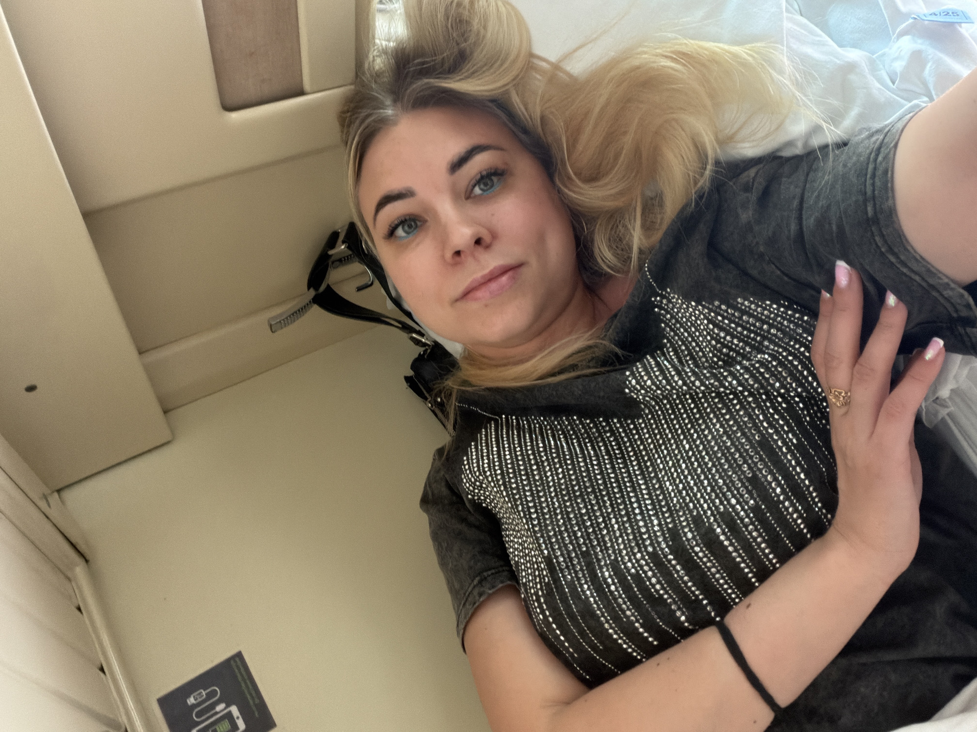 A woman lies on a bed in what appears to be a train compartment. Her head is positioned at the top of the frame, with her blonde hair styled in loose curls and waves. She is wearing a dark grey t-shirt with silver bead embellishments. Her hands are visible near the bottom of the frame.
The background shows the interior of the train compartment, including beige walls and a small utility panel with a USB charging symbol. It is not possible to pinpoint the exact location in Варламово, Russia from this image alone.