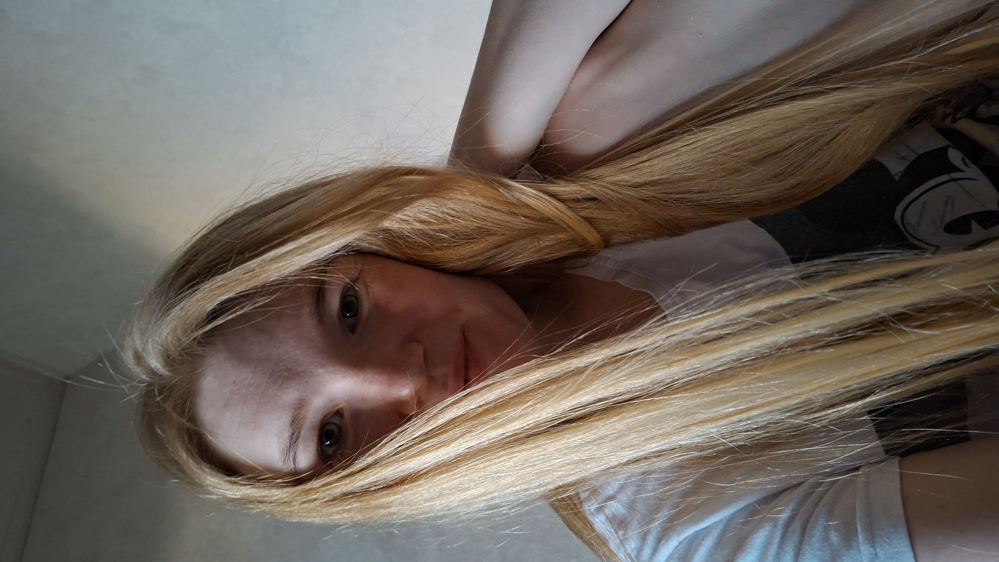 A young Caucasian woman with long blonde hair is taking a selfie in what appears to be a domestic setting. She is wearing a white t-shirt and has light skin with freckles. Her hair is partially covering her face. The background is a textured wall. There are no visible indications of the specific location within Большой Букор, Russia.