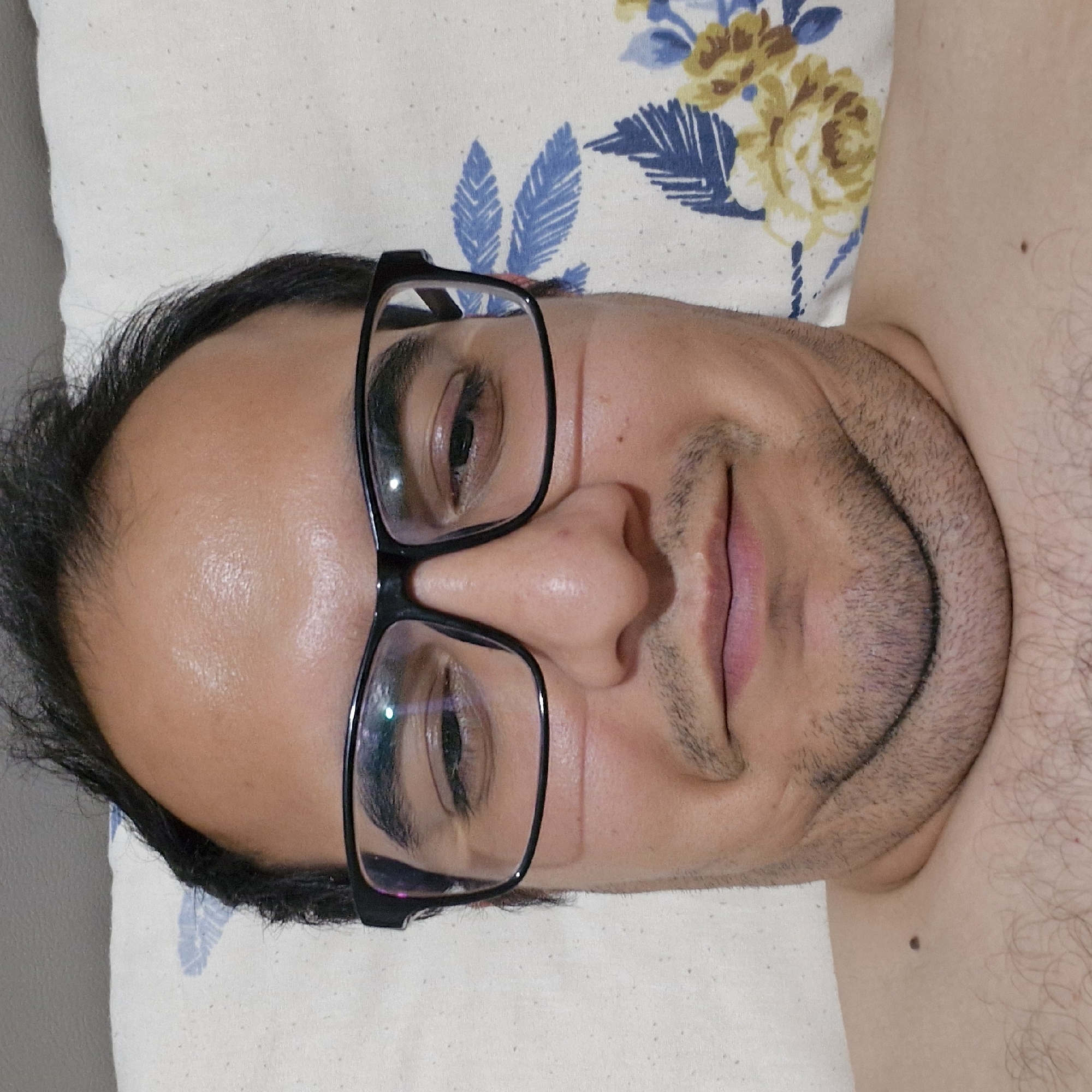 The image is a close-up, selfie-style photograph of a person's face, taken from a low angle. The subject is wearing black-rimmed glasses and has short, dark hair. The background appears to be a pillow with a floral pattern featuring blue leaves and yellow flowers. The subject's chin and neck are prominent in the lower portion of the frame, showing a rounded chin and visible facial hair. The lighting suggests an indoor setting, possibly a bedroom, with some reflections visible on the glasses. There is no indication of the city of Küçükçekmece, Turkey, or any urban environment.