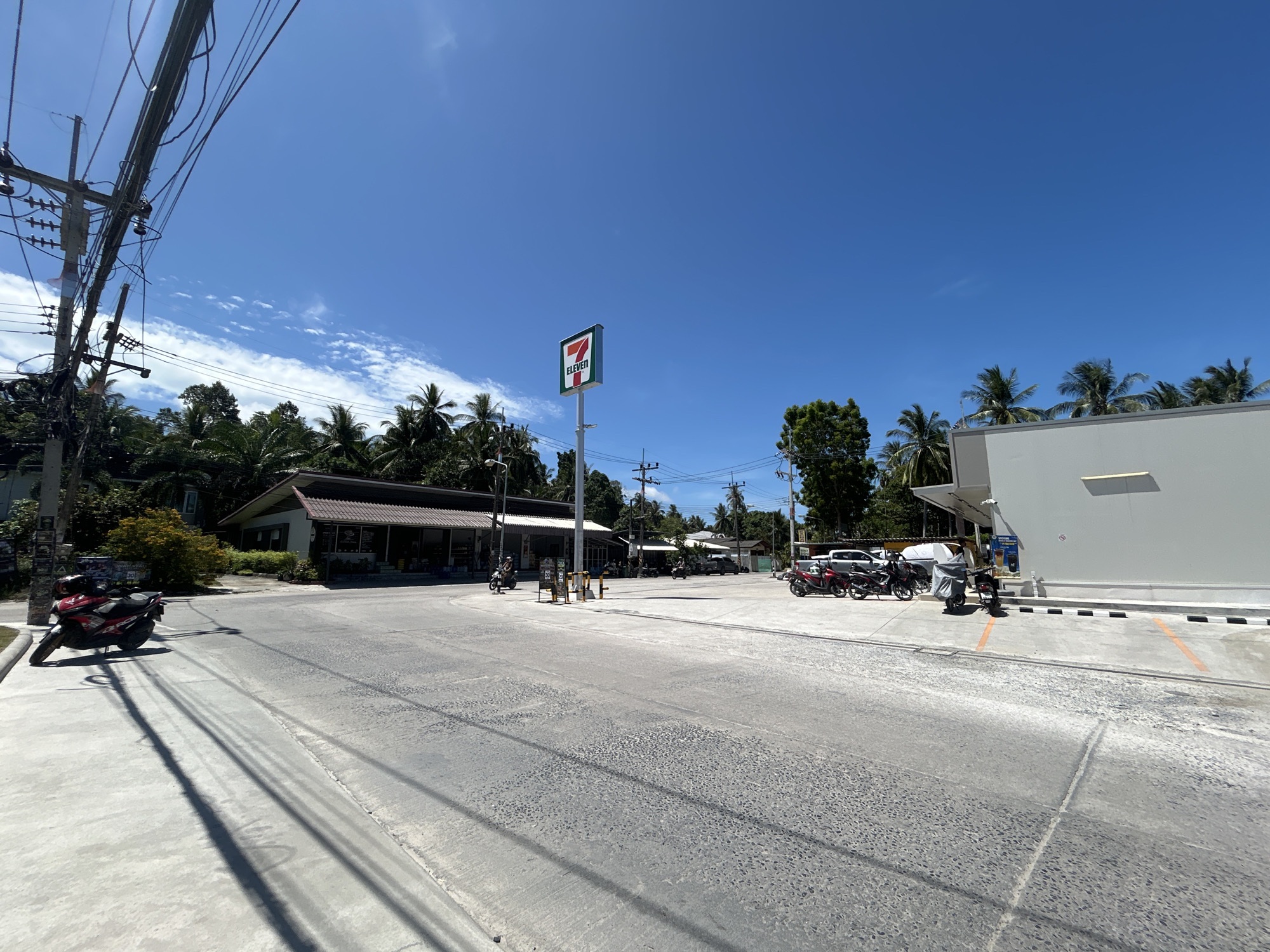 The image depicts a street scene in ตำบลเกาะพะงัน, Thailand, featuring a 7-Eleven store. The store is marked by its sign, centrally located. The area is paved with gray concrete, showing some wear. Several motorbikes are parked near the storefronts and along the roadside. Power lines are visible overhead, supported by utility poles. Trees and vegetation are in the background. The sky is blue with scattered clouds, indicating a sunny day. A red motorbike is parked on the left side of the road. Buildings and other businesses are present.